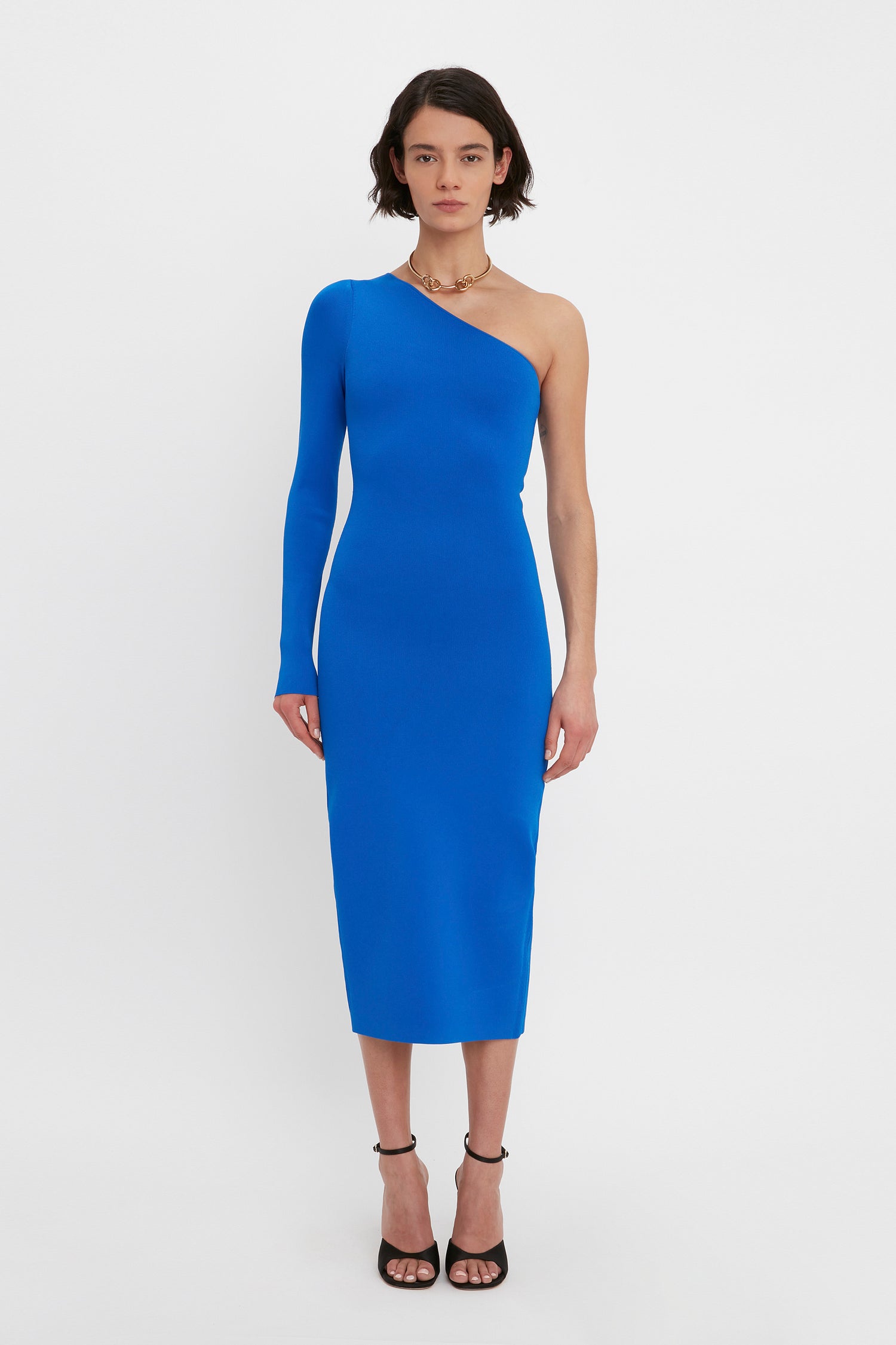 A person is standing against a white background, wearing the Victoria Beckham VB Body One Shoulder Midi Dress in Cobalt and black high-heeled sandals.