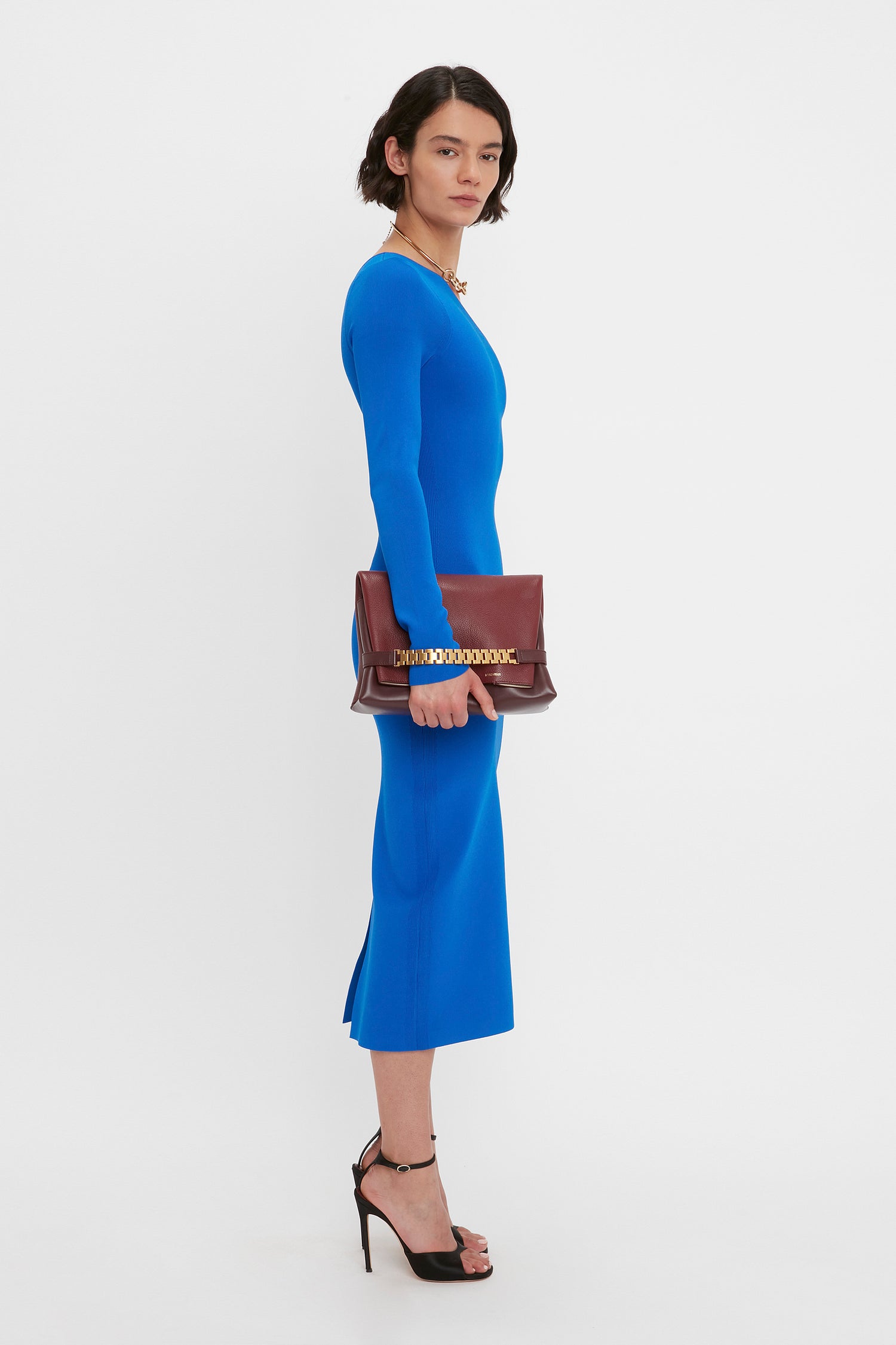 A person in a Victoria Beckham VB Body One Shoulder Midi Dress in Cobalt stands holding a brown clutch bag. They wear black high-heeled shoes and have a neutral expression.