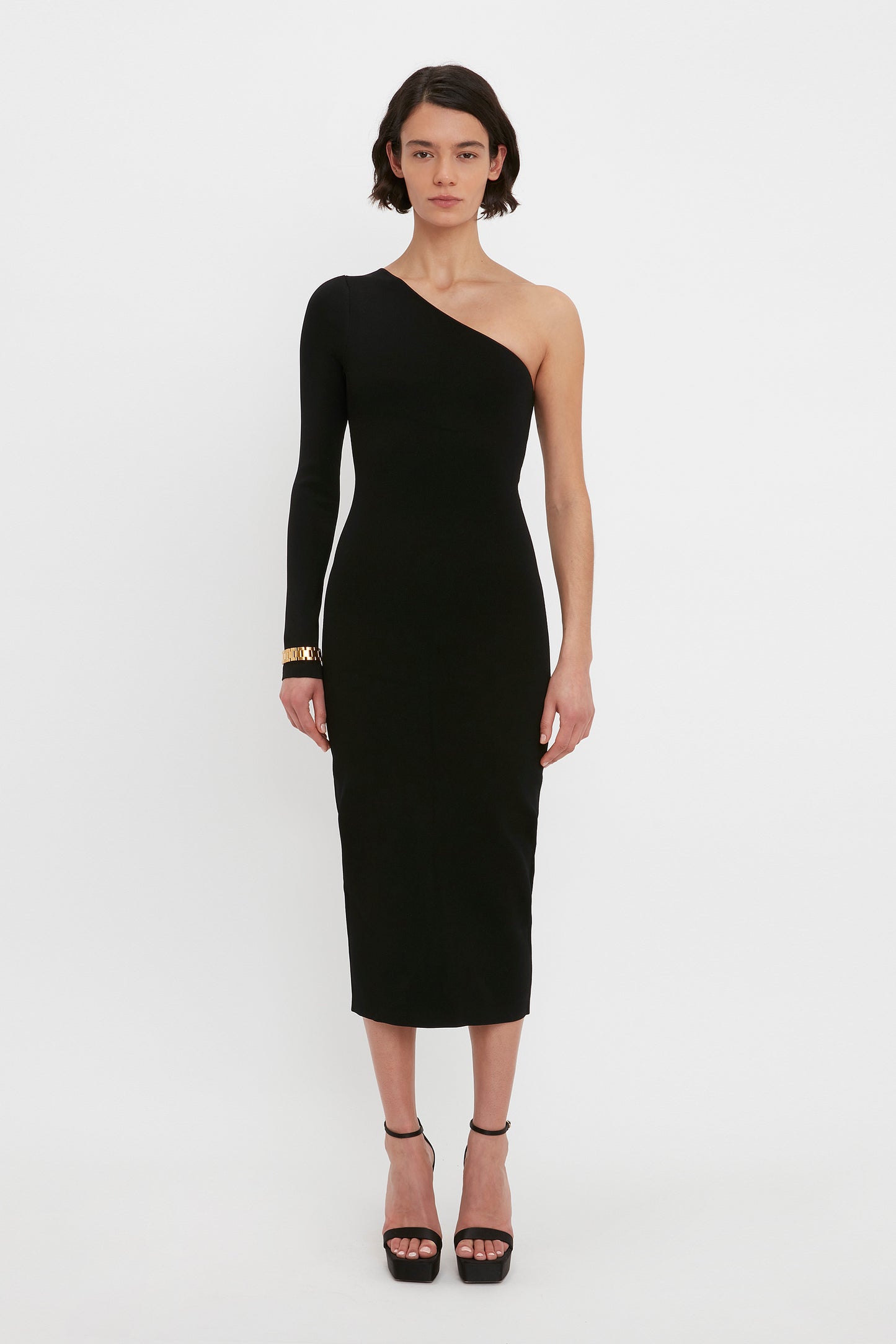 A person stands wearing the VB Body One Shoulder Midi Dress in Black by Victoria Beckham, paired with black high heels.