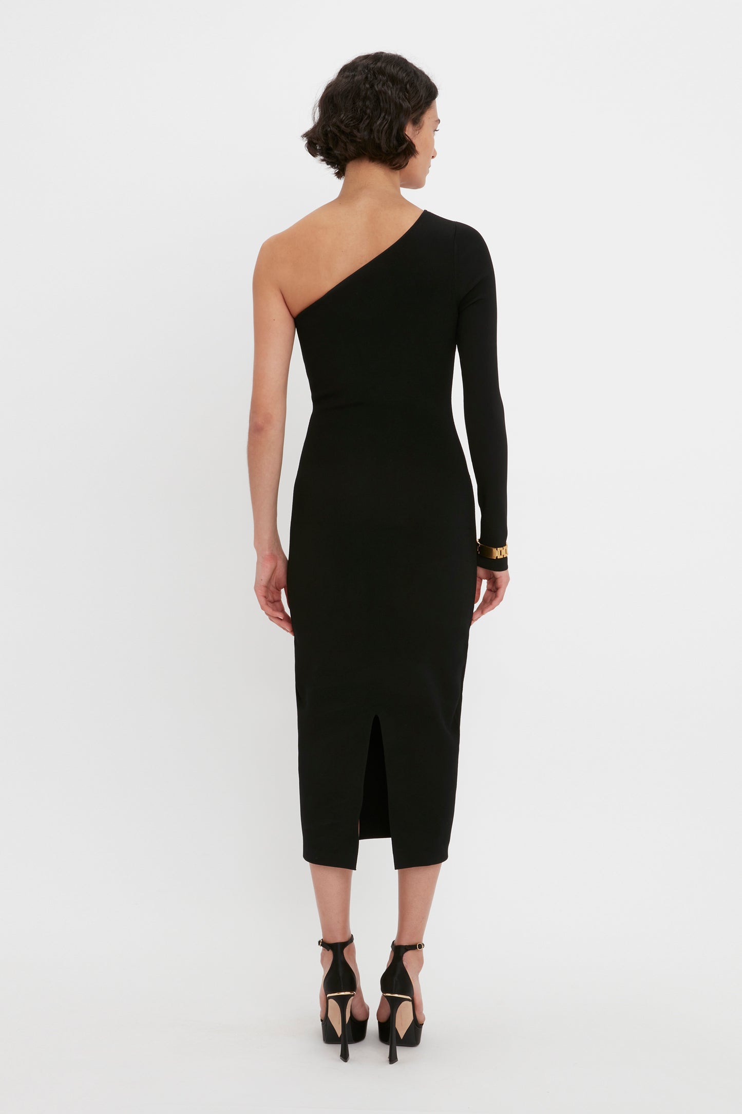 A person wearing the Victoria Beckham VB Body One Shoulder Midi Dress in Black, a form-fitting garment featuring a back slit, stands facing away.