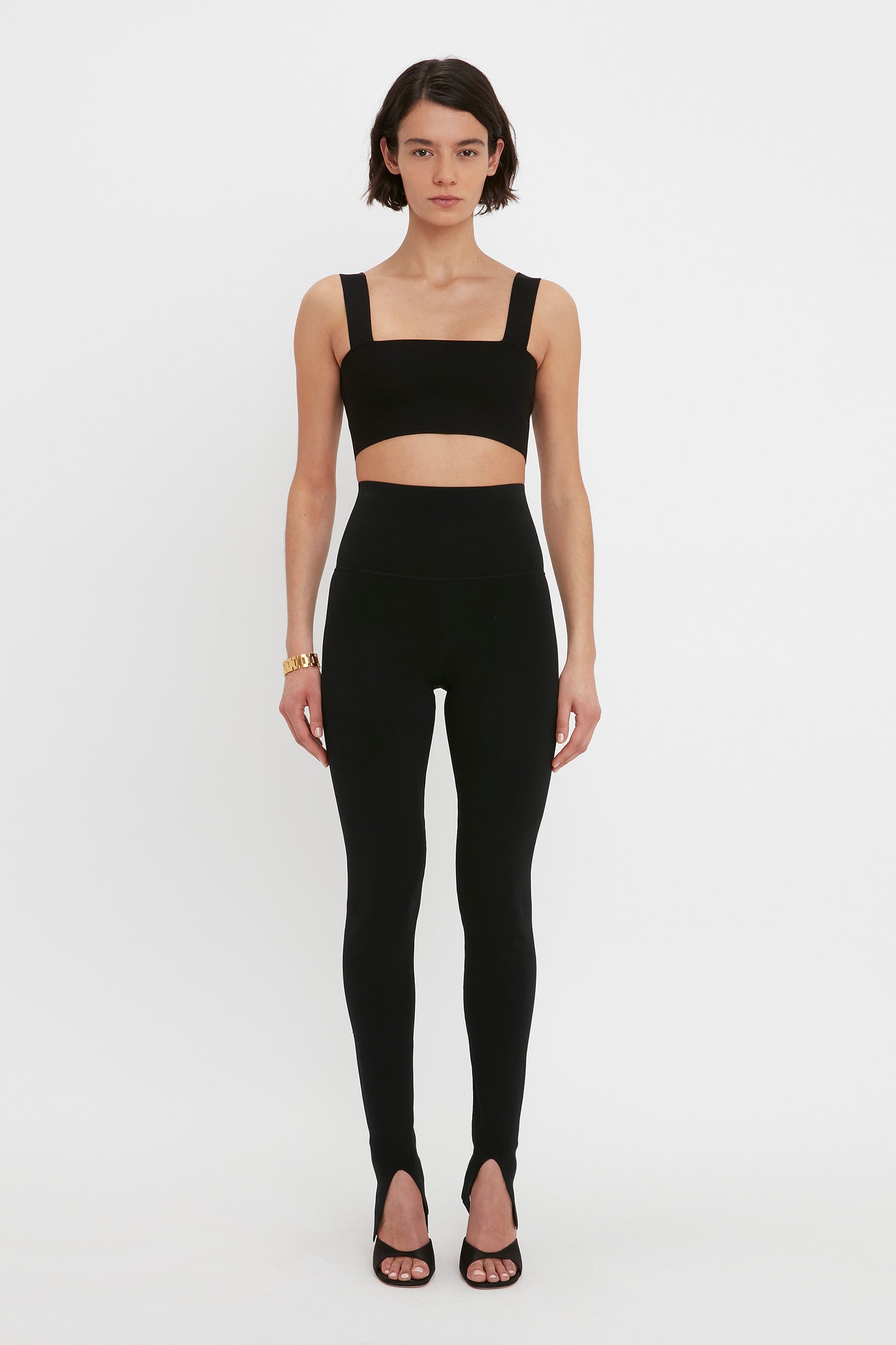 A woman with short dark hair is wearing the VB Body Strap Bandeau Top in Black by Victoria Beckham, high-waisted leggings, and black high-heeled shoes. She is standing against a plain white background.