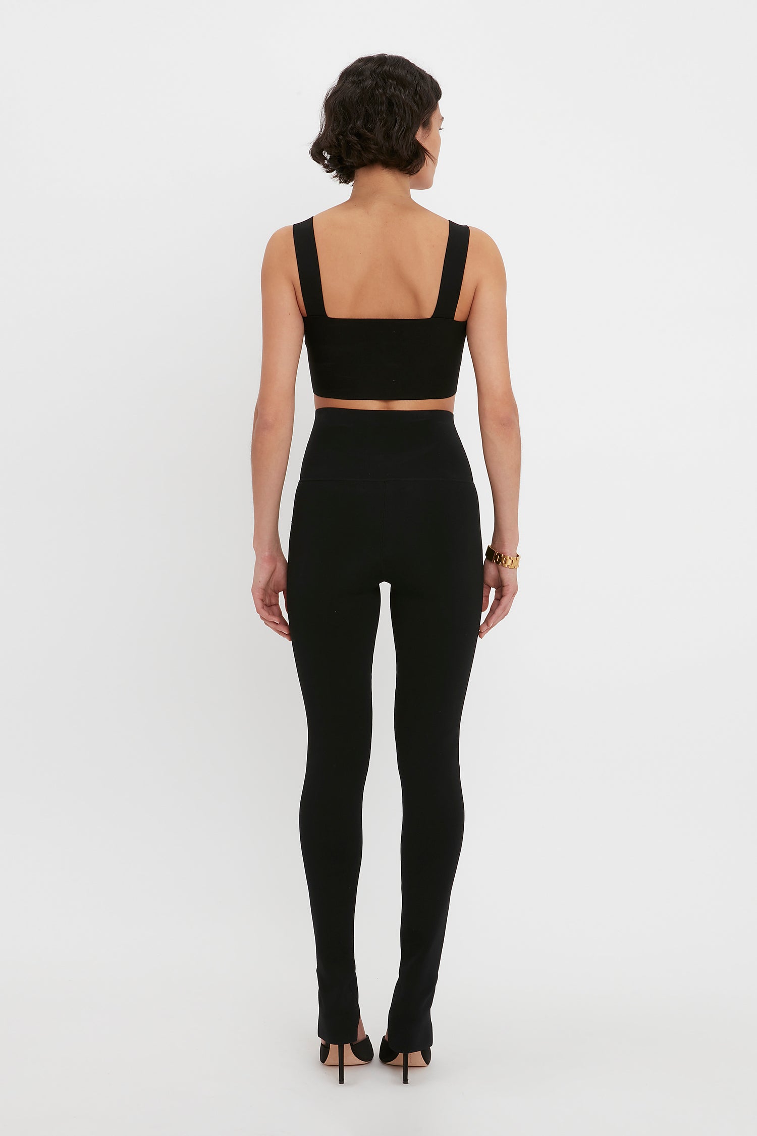 A person with short dark hair is seen from behind, wearing a VB Body Strap Bandeau Top in Black by Victoria Beckham, high-waisted black leggings, and black high heels.
