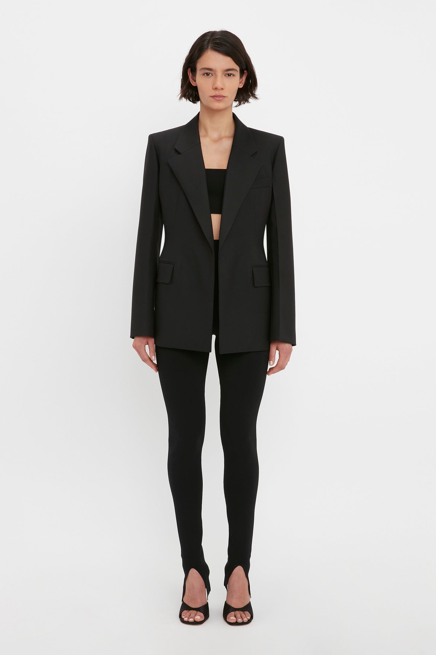 A person wearing a black blazer, the Victoria Beckham VB Body Split Front Leggings in Black, and black sandals stands against a plain white background.