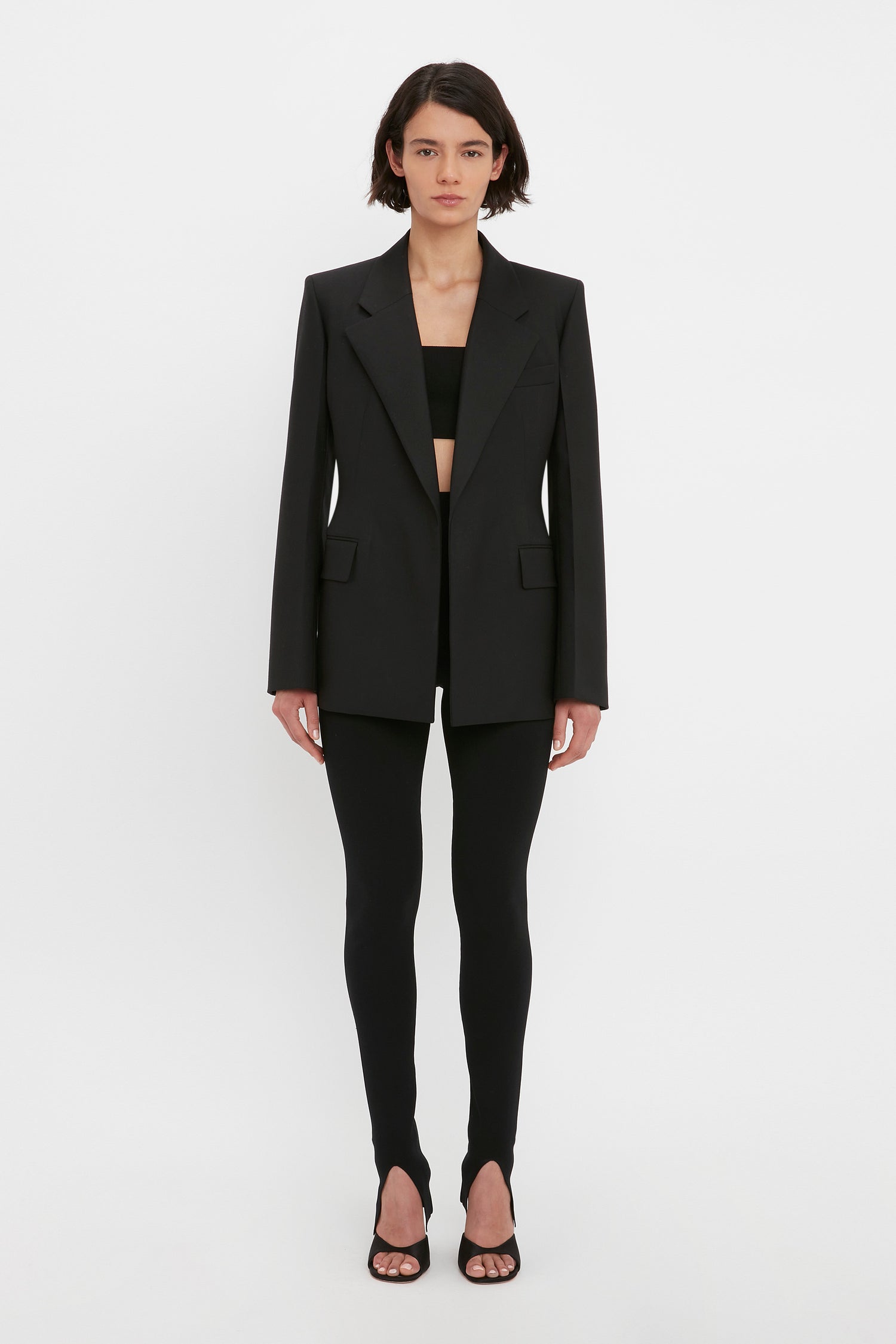 A person wearing a black blazer, the Victoria Beckham VB Body Split Front Leggings in Black, and black sandals stands against a plain white background.