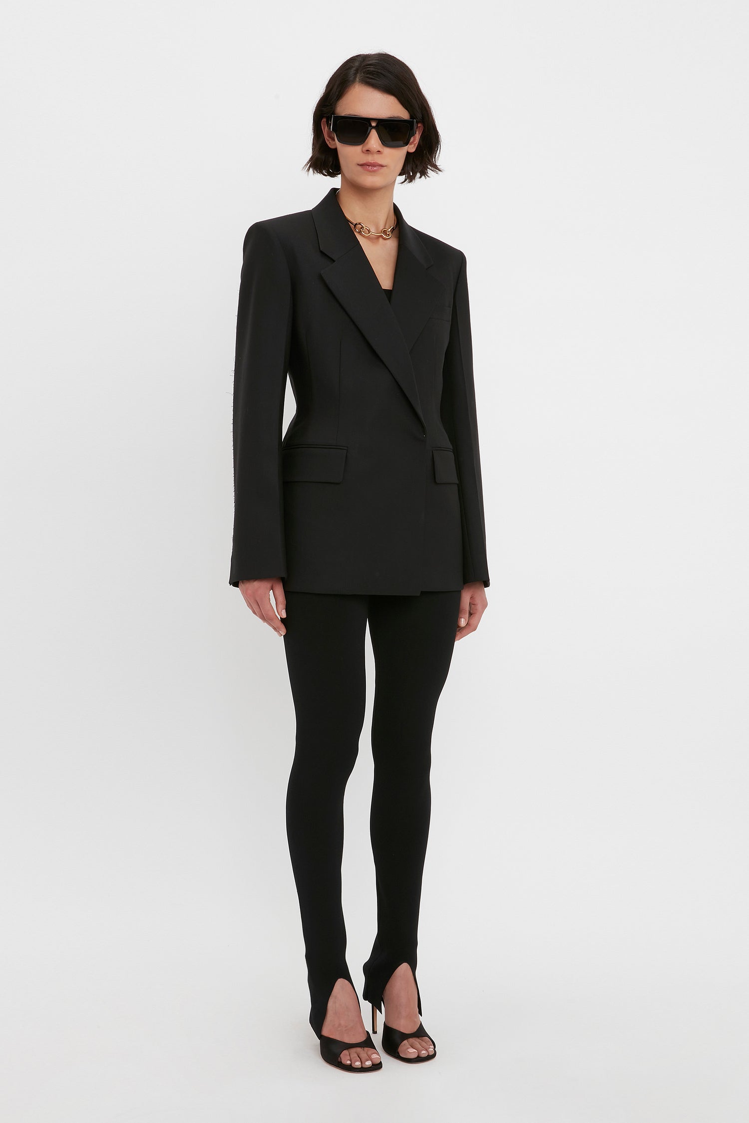 Person wearing a black blazer, VB Body Split Front Leggings in Black by Victoria Beckham, black sandals, sunglasses, and a gold chain necklace, standing against a plain white background.