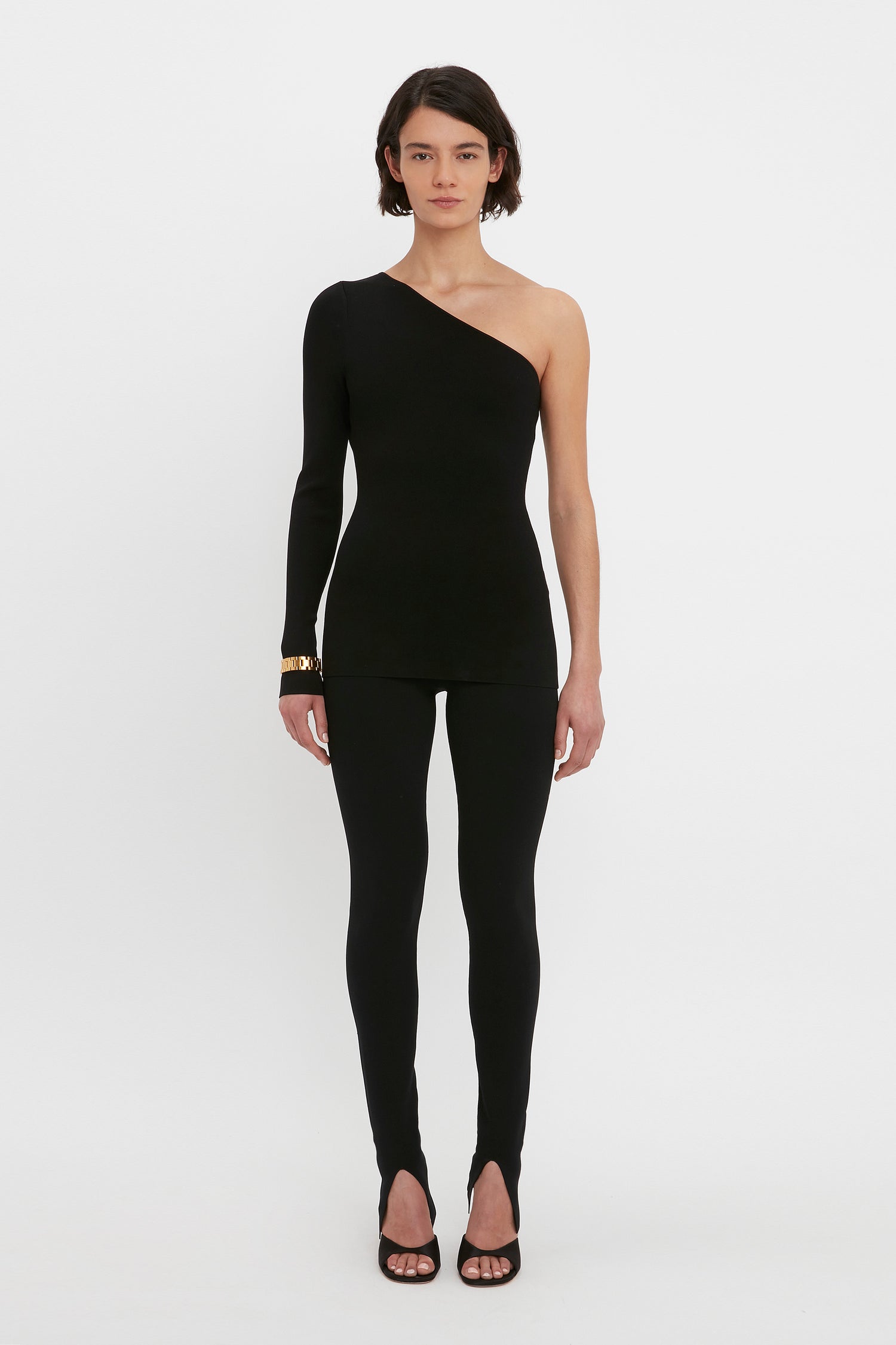A person stands against a white background wearing the VB Body One Shoulder Top in Black by Victoria Beckham along with matching black, fitted pants. They are also wearing black shoes with open toes.