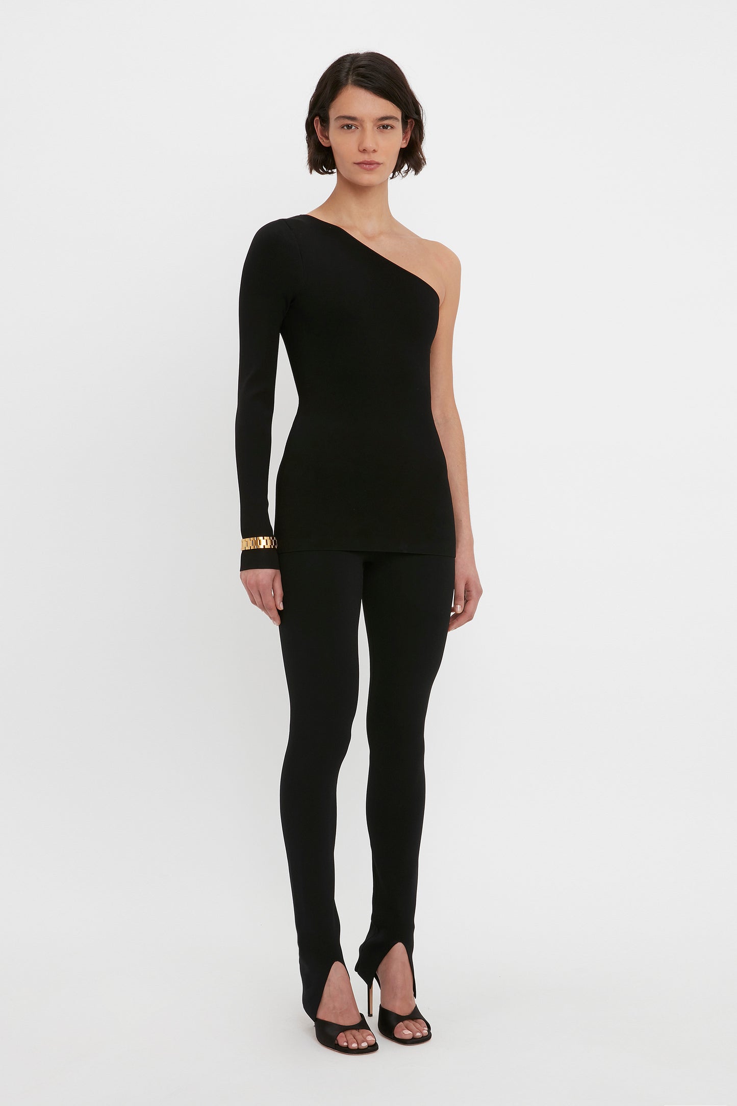 A person stands against a plain background, wearing the VB Body One Shoulder Top in Black by Victoria Beckham and matching black leggings with split hems. They are also wearing black high-heeled shoes and a gold bracelet on one wrist.