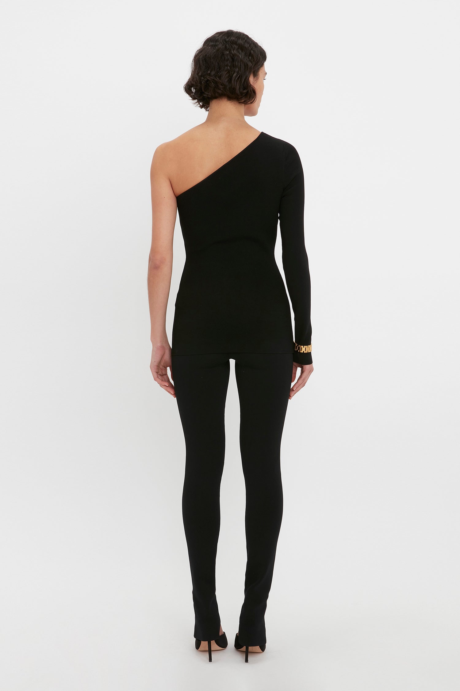 A person stands against a plain background wearing the VB Body One Shoulder Top in Black by Victoria Beckham, showcasing a bare left shoulder and paired with full-length pants, viewed from the back.
