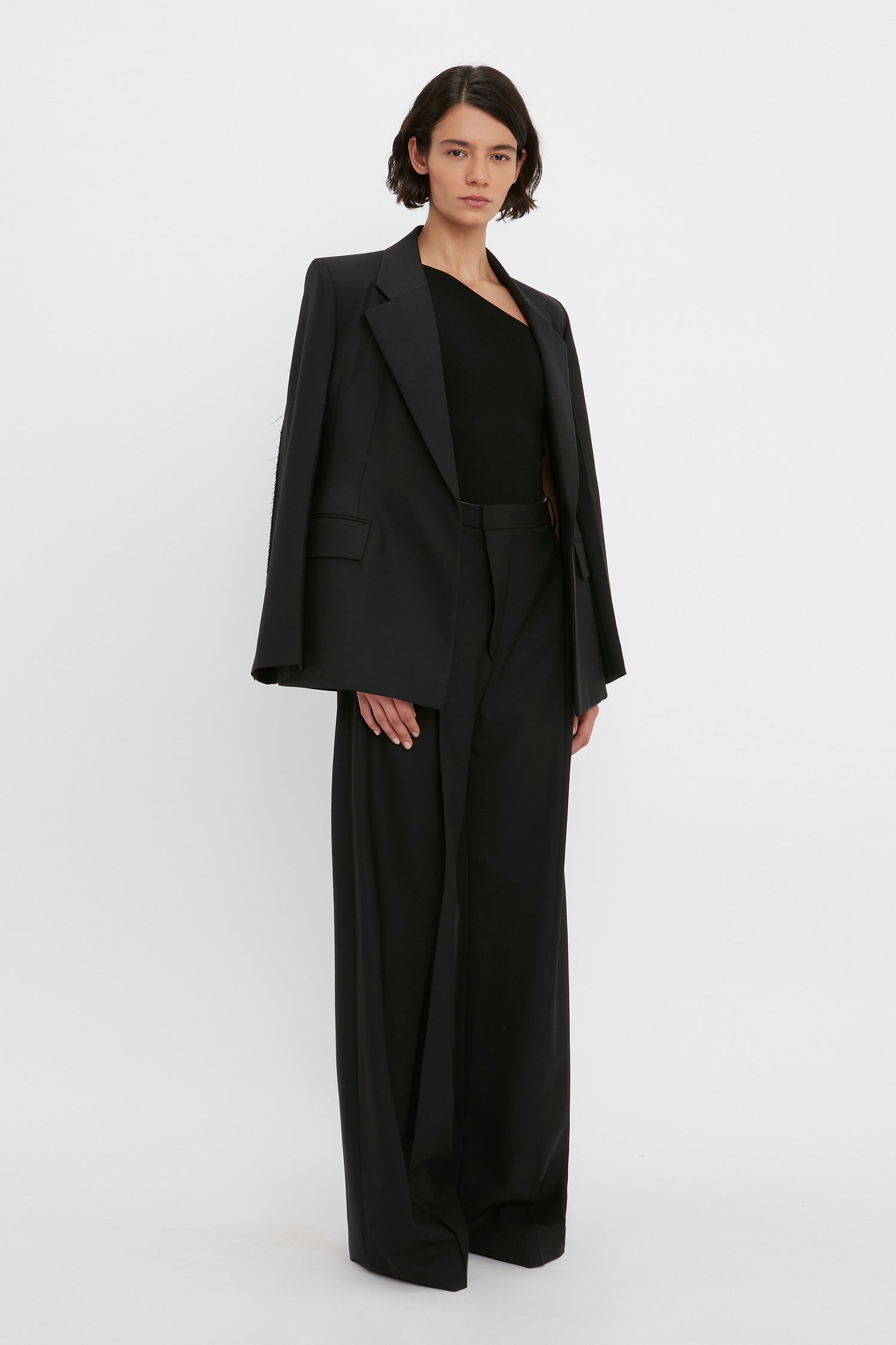 A person wearing a black blazer, the VB Body One Shoulder Top in Black by Victoria Beckham, and black wide-leg trousers stands against a plain white background.