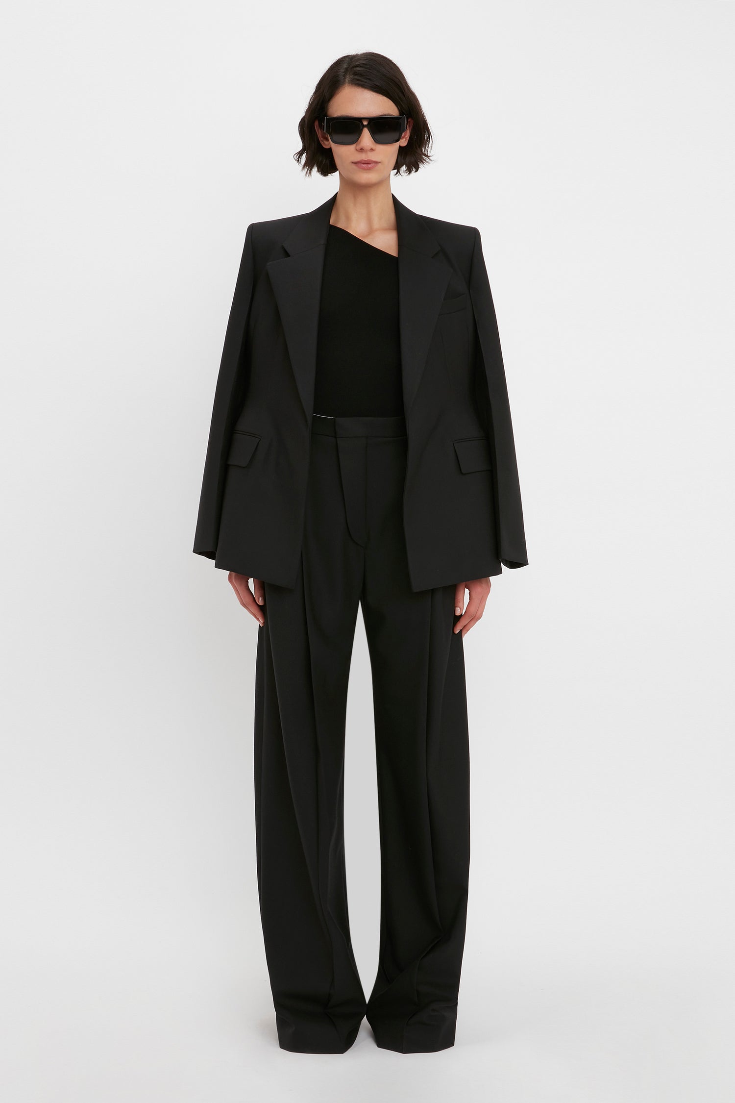 A confident individual stands against a plain white background, wearing an all-black outfit that includes the Victoria Beckham VB Body One Shoulder Top in Black, a blazer draped over their shoulders, wide-legged trousers, and sunglasses.