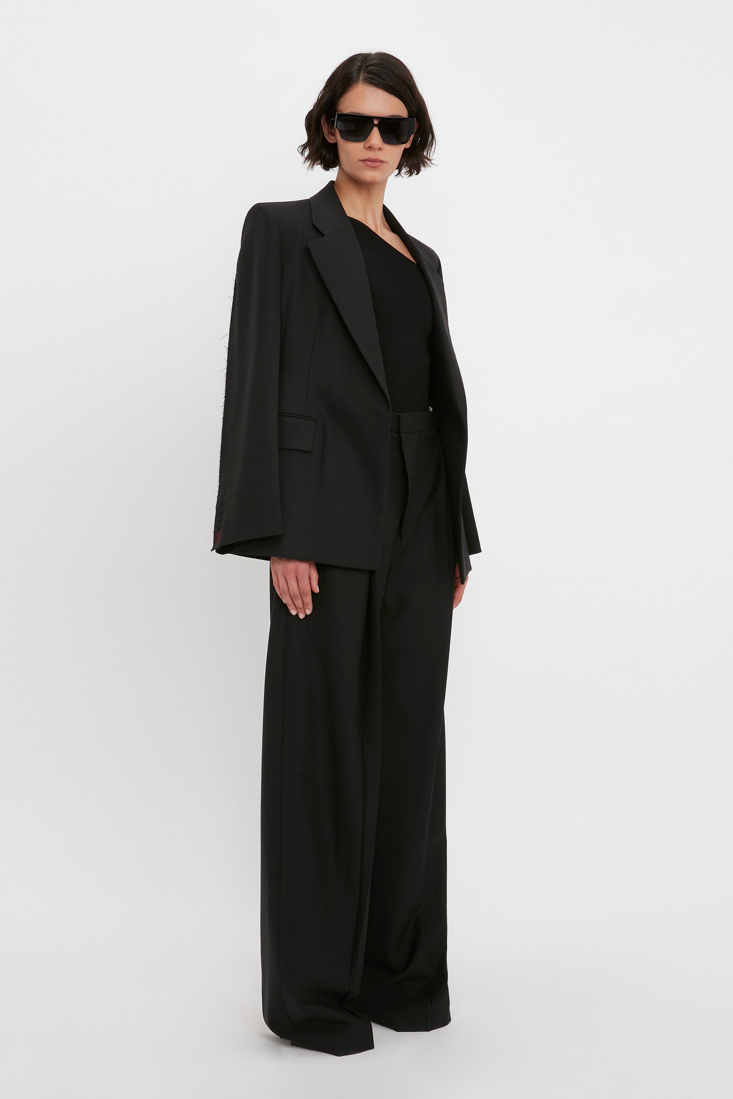 A person wearing a black suit, the VB Body One Shoulder Top in Black by Victoria Beckham, and sunglasses stands against a plain white background.
