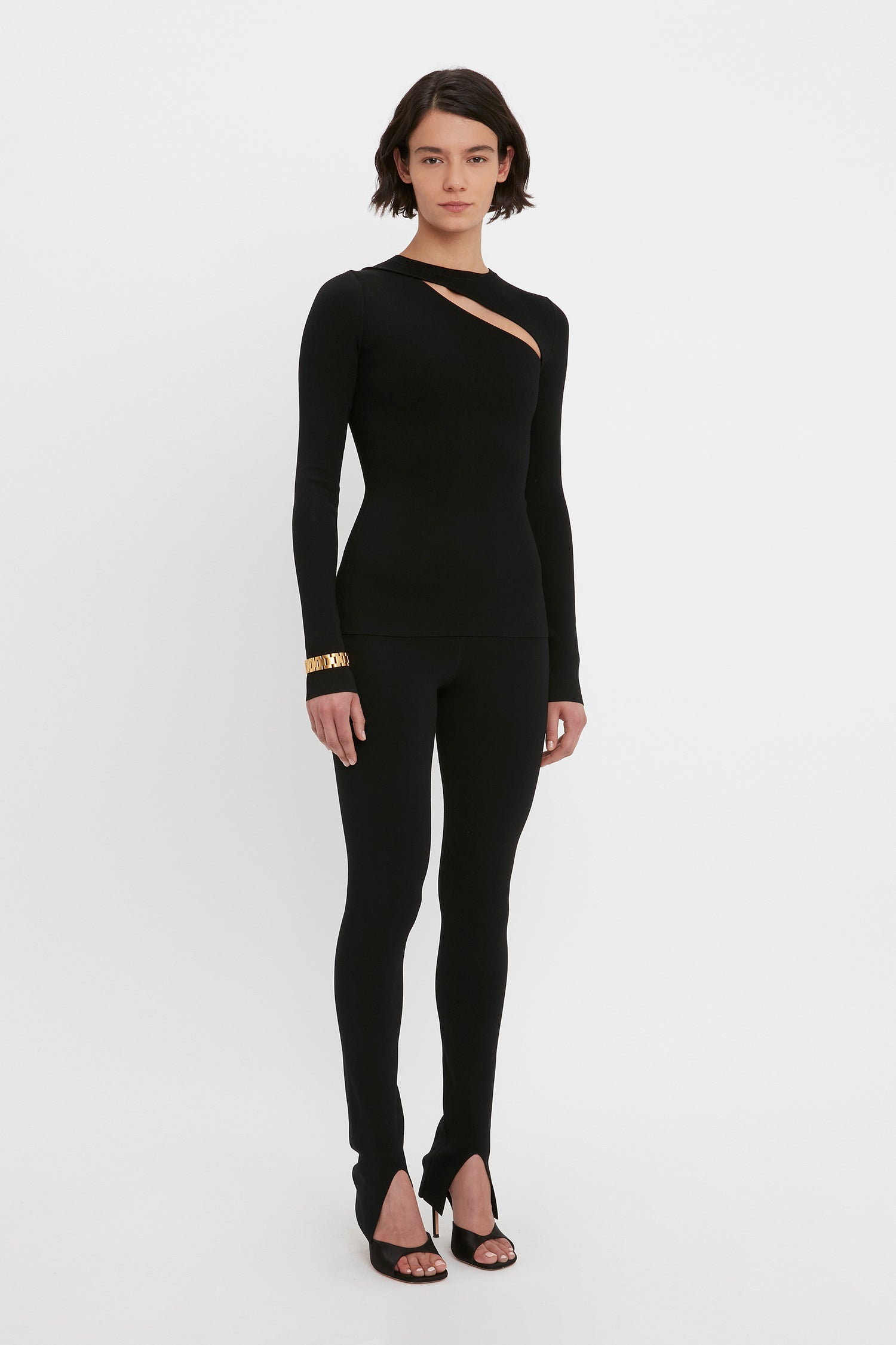 A person stands against a white background wearing a black, long-sleeve VB Body Front Slash Crew Neck Top by Victoria Beckham. The outfit is form-fitting with a diagonal cutout on the chest. They have short, dark hair, and are accessorized with black heels and a gold bracelet.