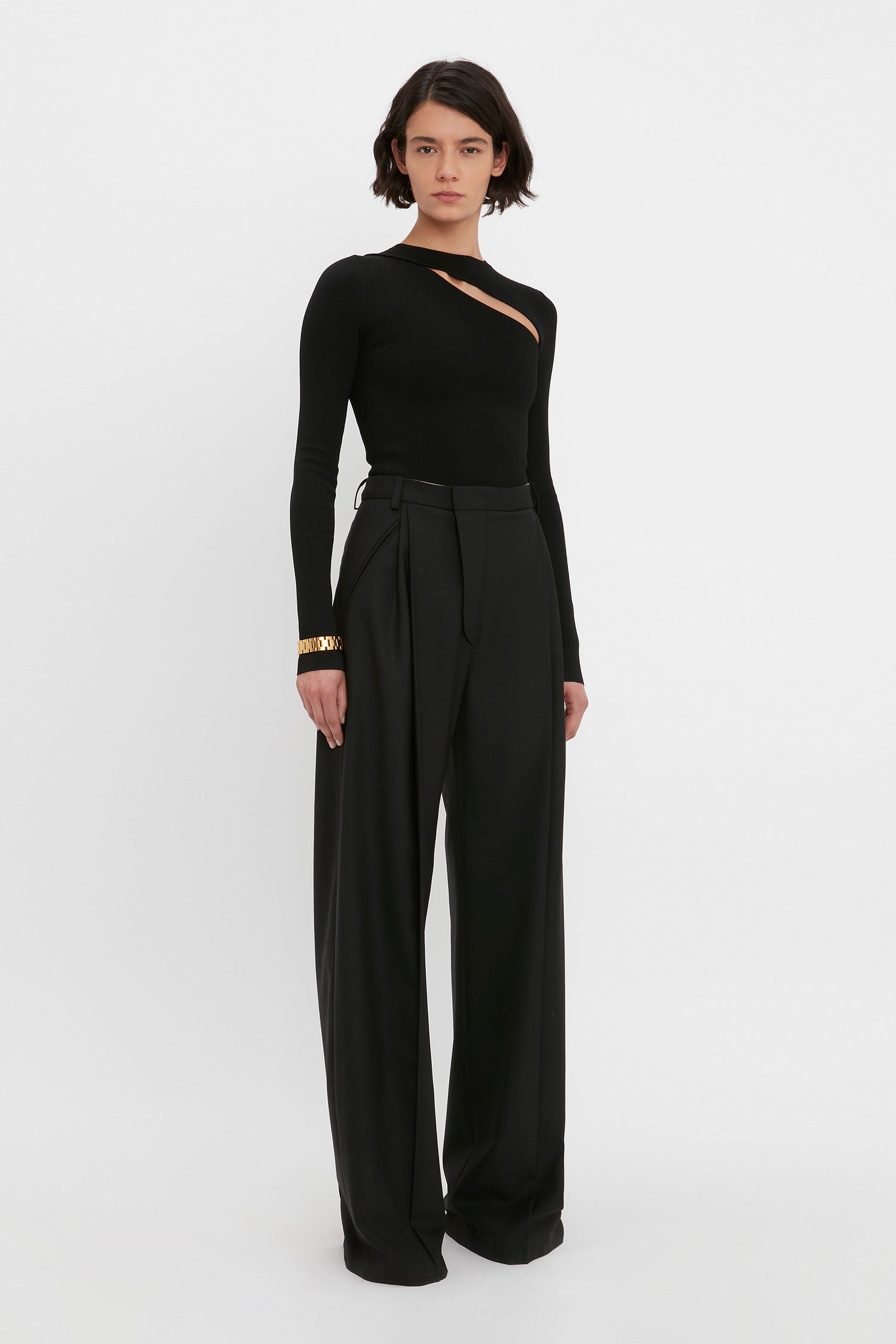 Standing against a plain white background, a person wears the VB Body Front Slash Crew Neck Top in Black by Victoria Beckham, paired with high-waisted, wide-leg black trousers.