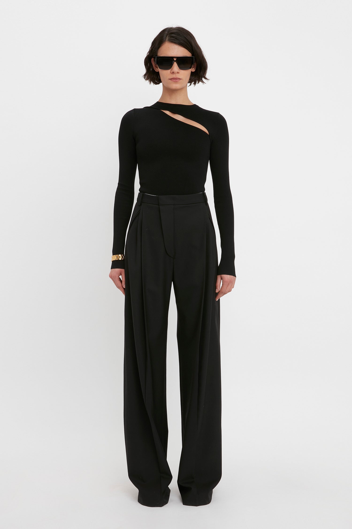 A person stands against a white background wearing the VB Body Front Slash Crew Neck Top in black by Victoria Beckham, high-waisted wide-leg black trousers, and dark sunglasses. Their arms hang straight down by their sides.