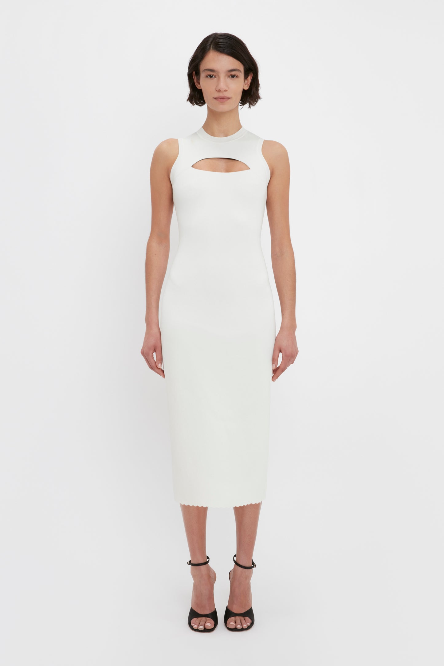 A woman stands facing forward wearing the VB Body Slash Front Midi Dress in White by Victoria Beckham, a sleeveless, white, mid-calf dress with a front cut-out detail. She is also wearing black open-toe high heels.