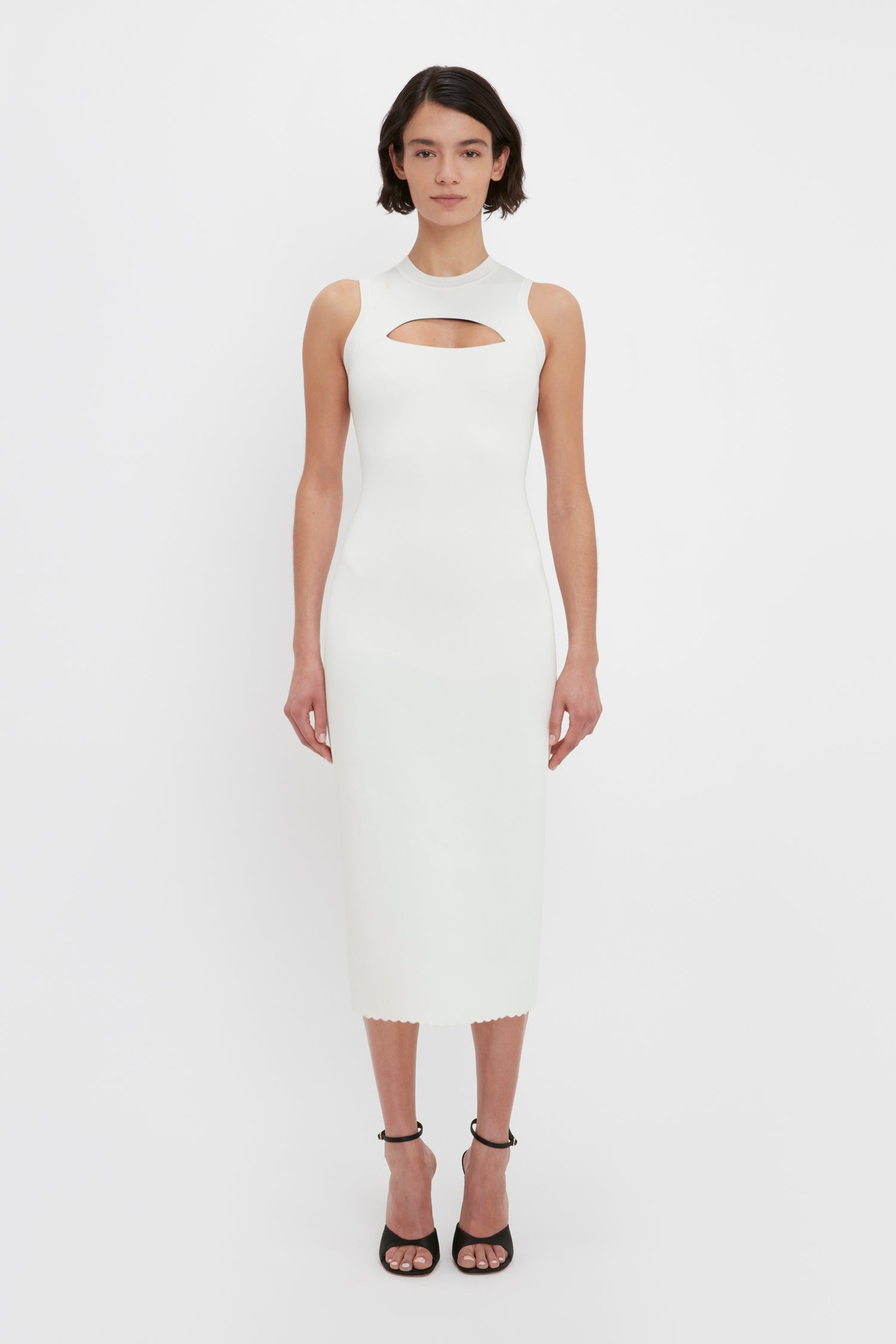 A woman stands facing forward wearing the VB Body Slash Front Midi Dress in White by Victoria Beckham, a sleeveless, white, mid-calf dress with a front cut-out detail. She is also wearing black open-toe high heels.