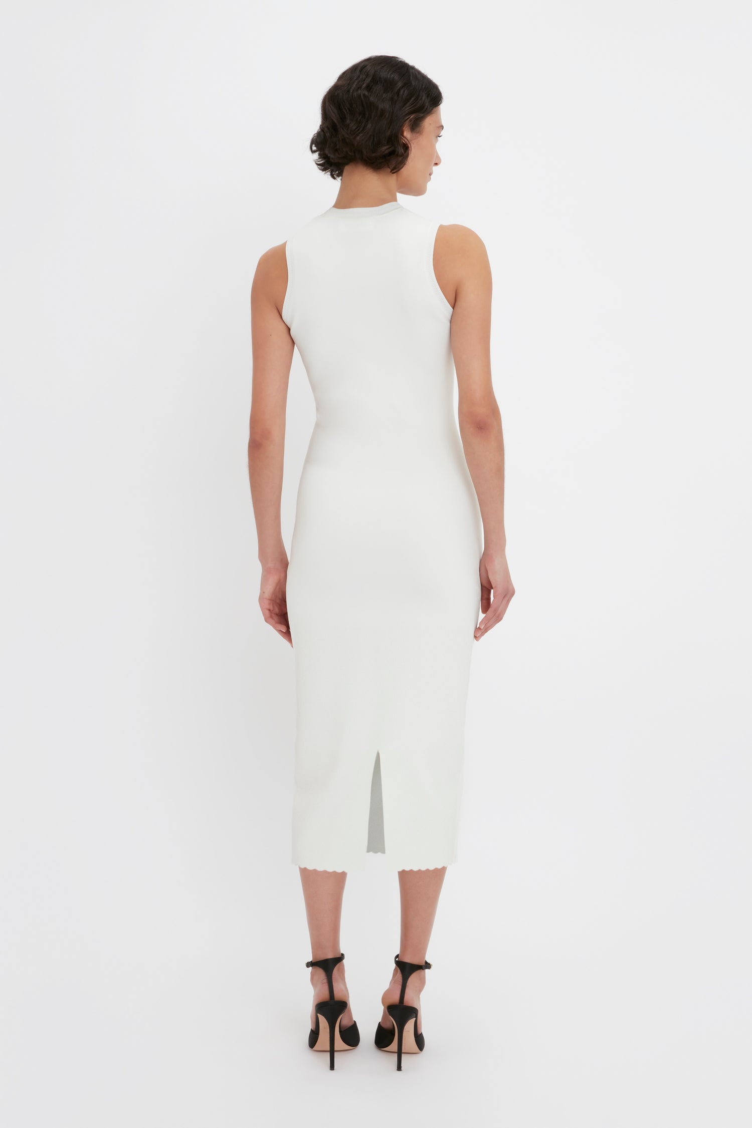 A person with short dark hair is seen from the back, wearing a sleeveless, form-fitting VB Body Slash Front Midi Dress in White by Victoria Beckham and black high heels.