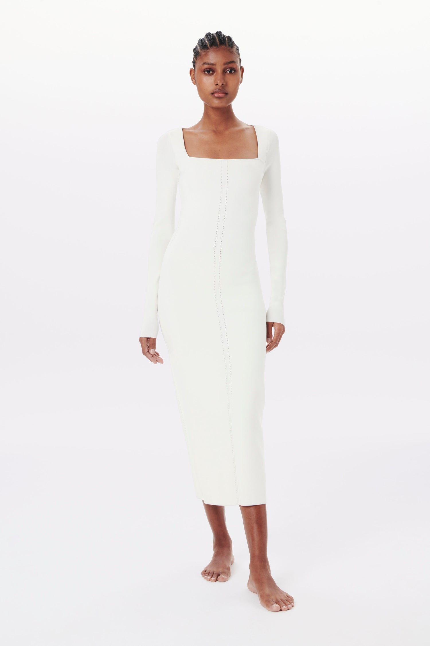 A person wearing the VB BODY 1 dress by Victoria Beckham, a white, long-sleeved, square-neck, form-fitting midi dress, walking barefoot in a brightly lit, white studio.