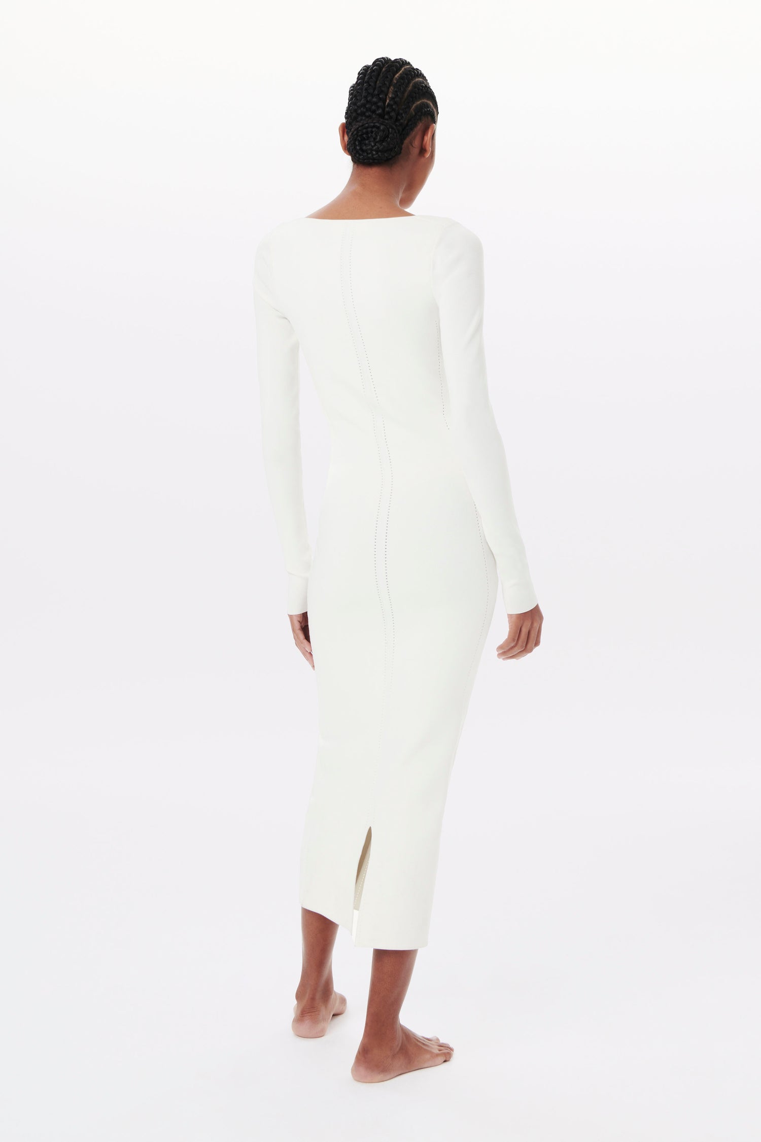 Revised Sentence: Rear view of a person with braided hair wearing the Victoria Beckham VB BODY 1, a long-sleeved, white, fitted dress. They are barefoot and standing against a plain white background.