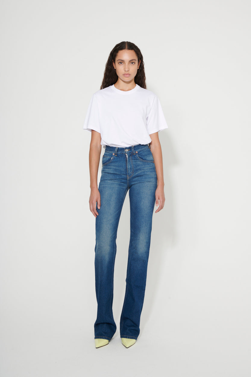A woman stands against a plain white background, wearing the Victoria Beckham MAW22 4 white T-shirt, high-waisted blue jeans, and light-colored shoes.
