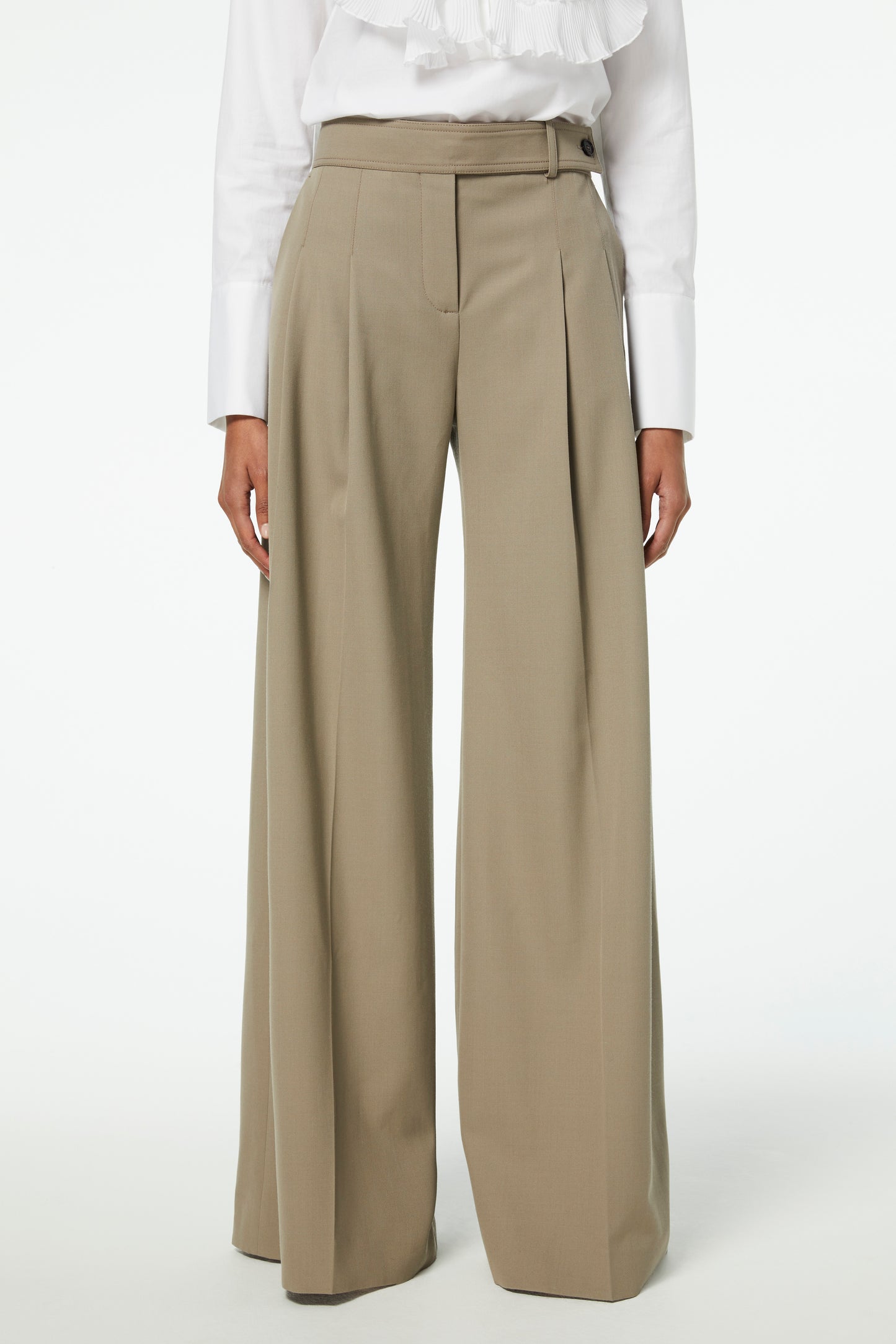 A person is seen wearing Victoria Beckham's Wide-leg Trousers in Clay, made of high-waisted wool gabardine, paired with a crisp white blouse. The image captures them from shoulders to feet against a pristine white background.