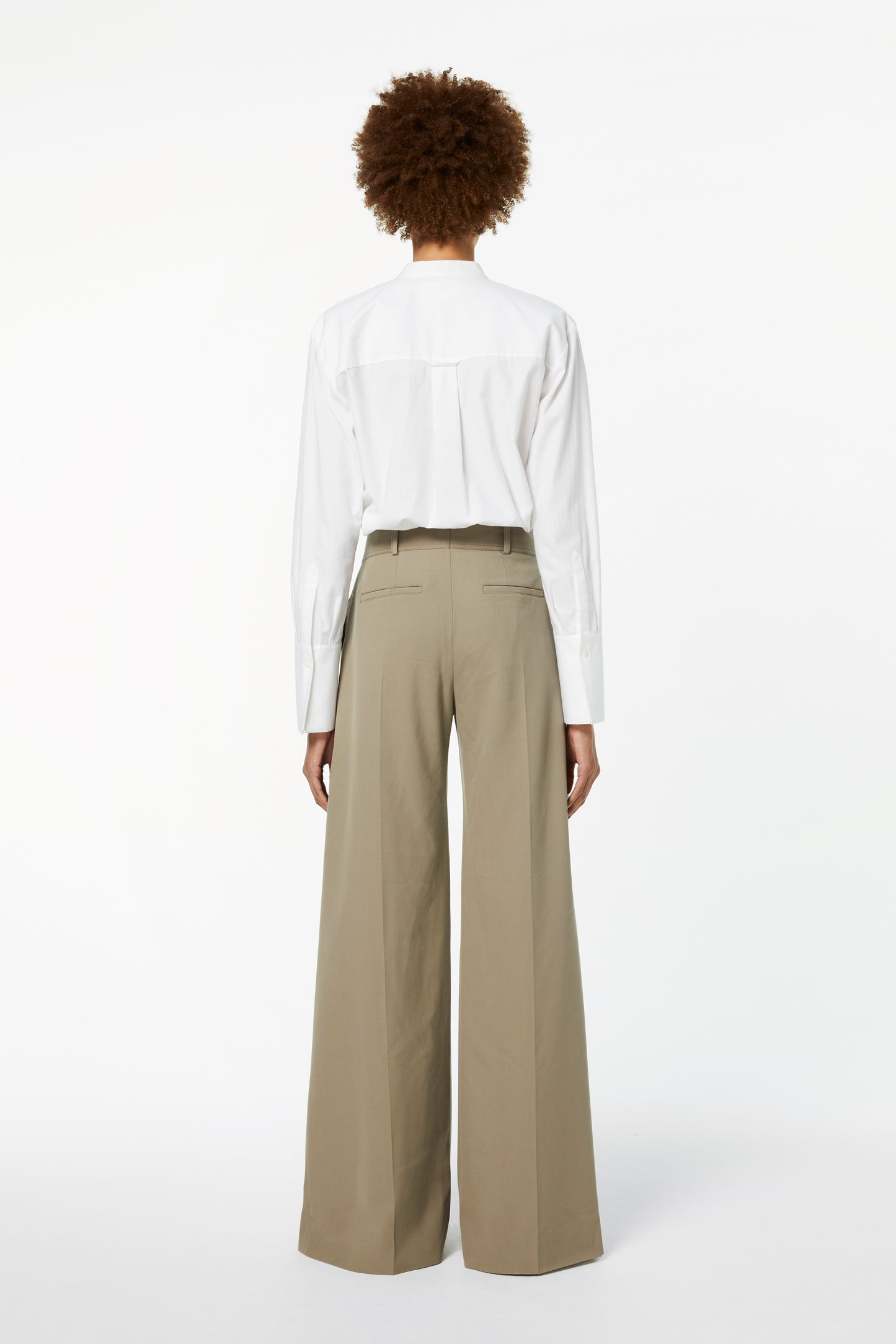 A person with curly hair is seen from behind wearing a white long-sleeve shirt and Victoria Beckham's Wide-leg Trousers in Clay.