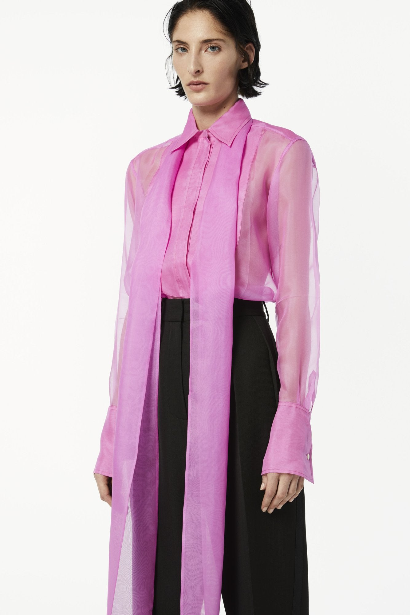 A person stands wearing a sheer hot pink blouse with a long scarf detail, black trousers, and a Victoria Beckham bundletest T-shirt draped stylishly over their shoulder, looking directly at the camera.