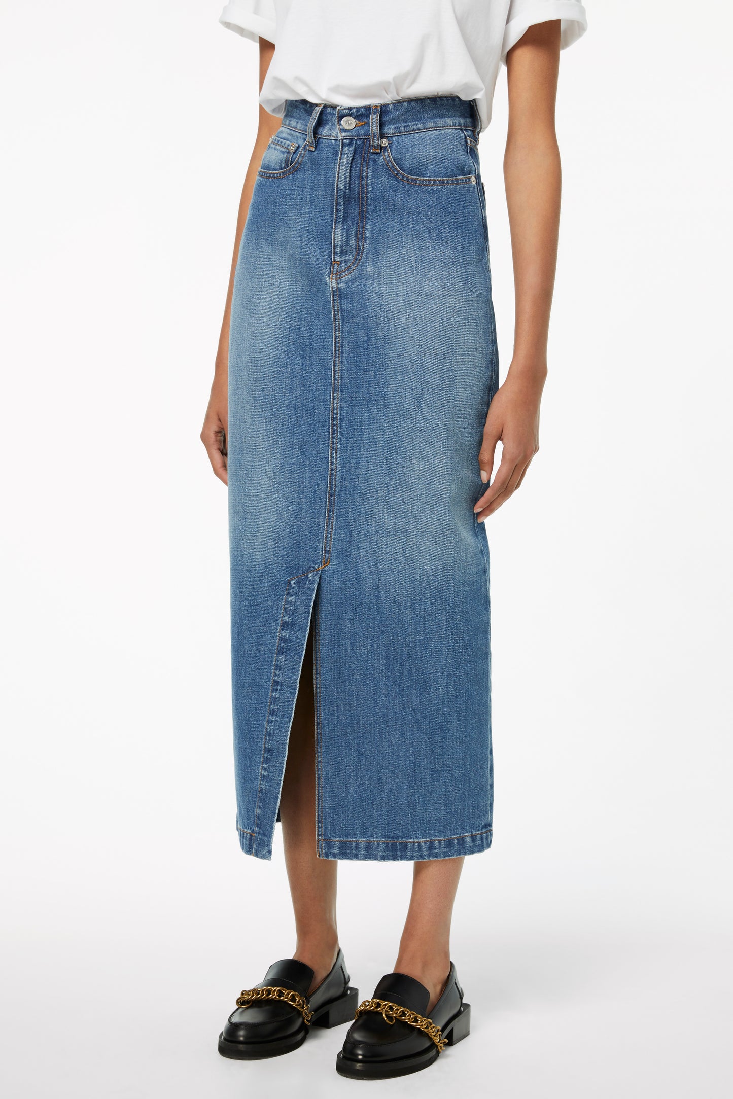 A person wearing a white shirt in a transitional style, paired with Victoria Beckham's Front Split Denim Midi Skirt in Fresh Blue and black loafers adorned with gold chain detail.