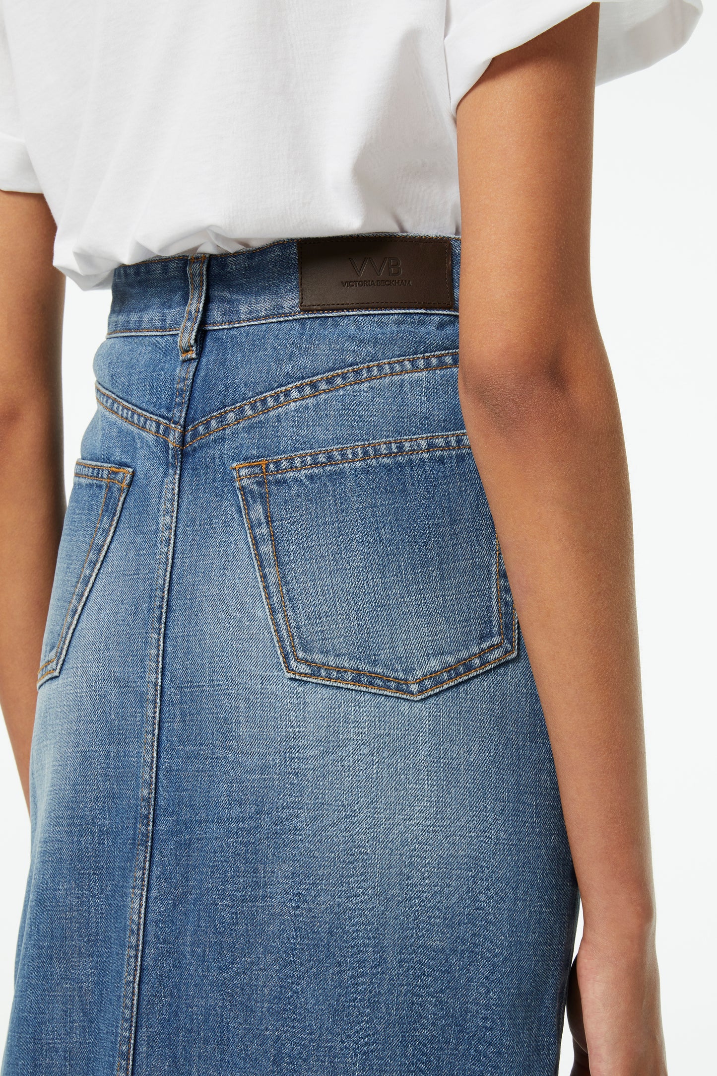 A woman showcases a transitional style in a Victoria Beckham Front Split Denim Midi Skirt in Fresh Blue with visible back pockets and a brown label, paired elegantly with a crisp white shirt.