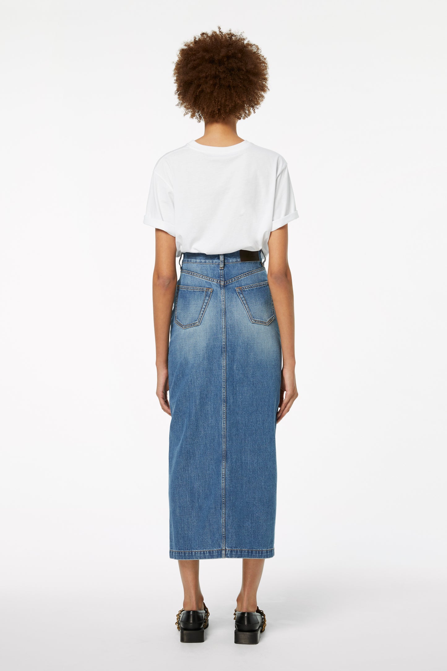 A person with curly hair stands facing away, wearing a white T-shirt, a Victoria Beckham Front Split Denim Midi Skirt in Fresh Blue, and black shoes, effortlessly embodying transitional style.