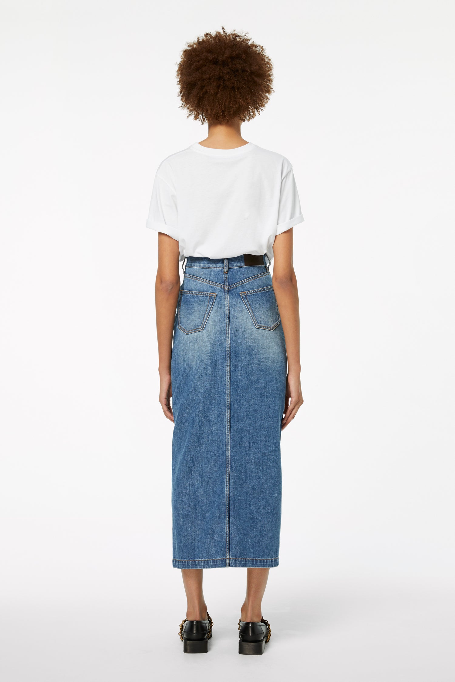 A person with curly hair stands facing away, wearing a white T-shirt, a Victoria Beckham Front Split Denim Midi Skirt in Fresh Blue, and black shoes, effortlessly embodying transitional style.