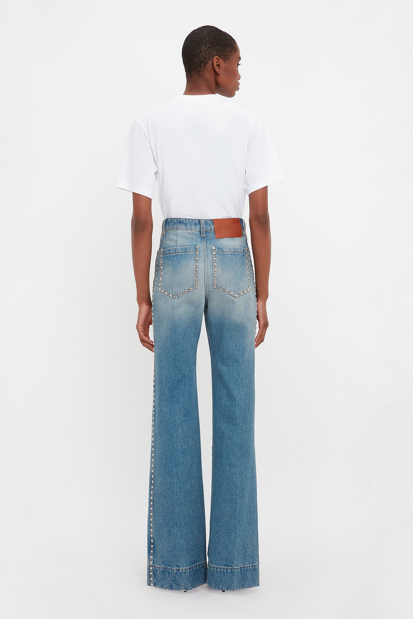 Facing away from the camera against a plain background, a person wears a white T-shirt paired with Victoria Beckham's BAO TEST R - Alina High Waisted Jean in Vintage Wash, featuring studs.