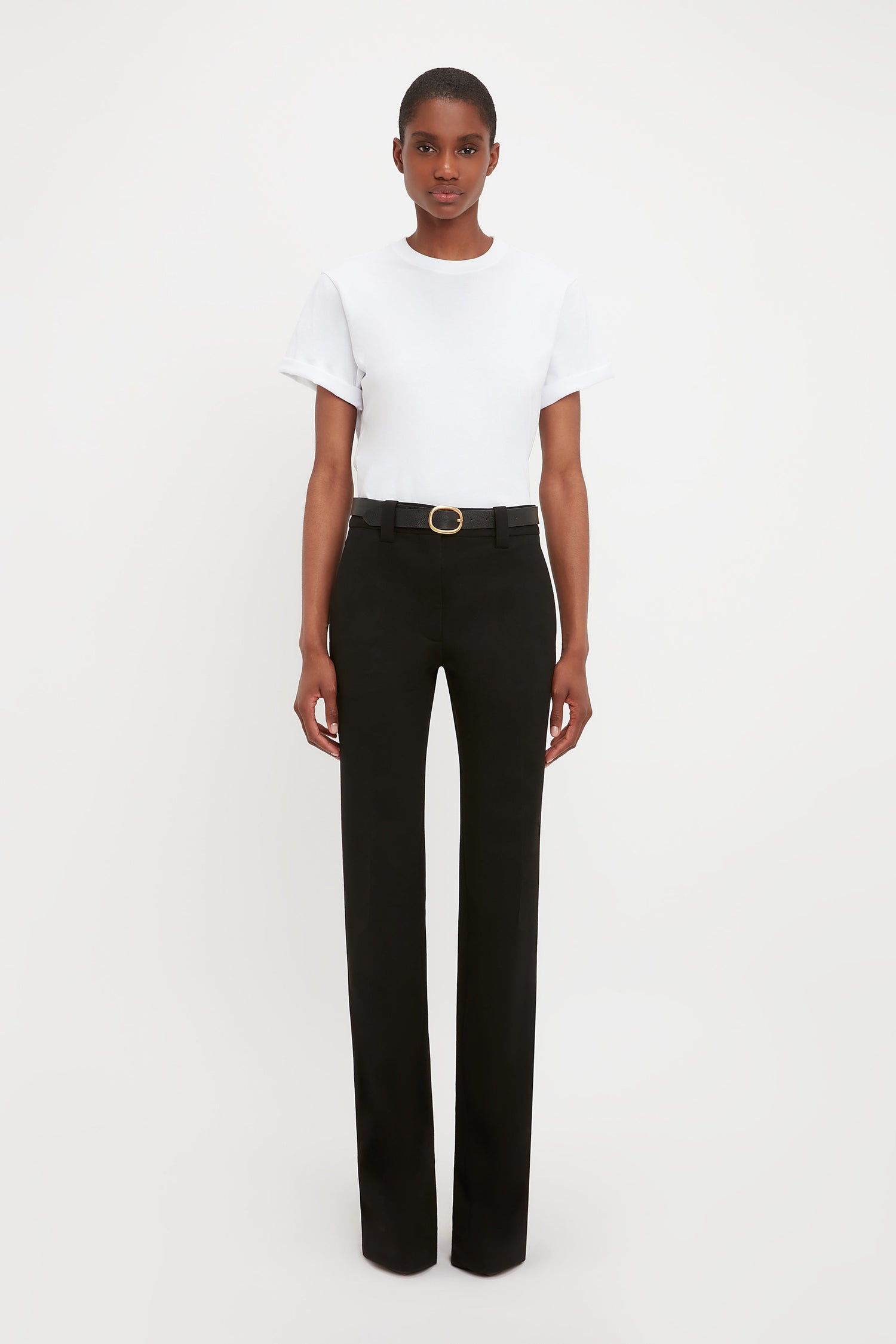 A person stands against a plain white background wearing a white short-sleeve t-shirt, Victoria Beckham Tailored Straight Leg Trouser in Black with a belt, and black shoes.