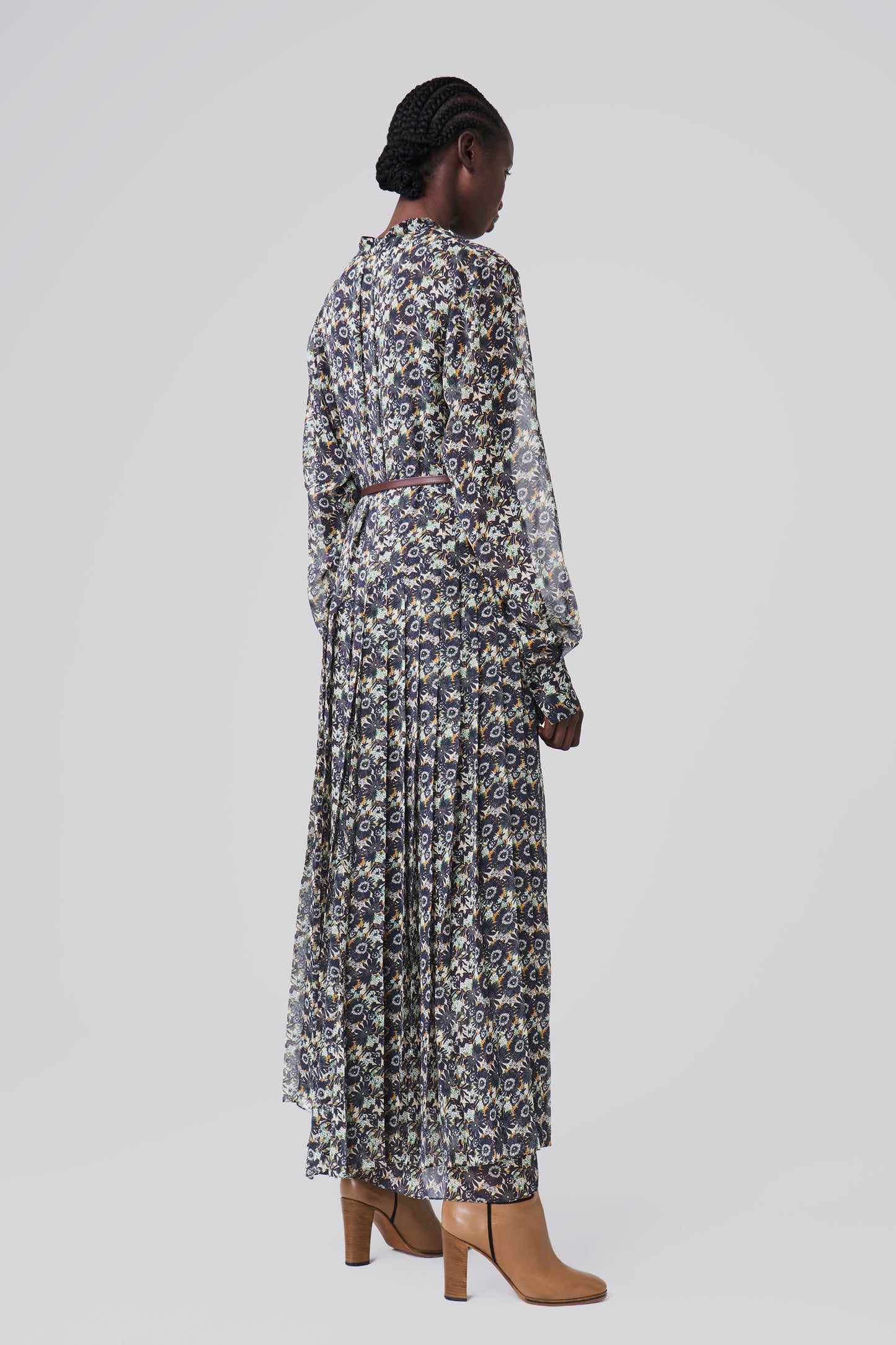 A woman in a pleated dress featuring a navy floral print from Victoria Beckham and brown ankle boots stands with her back to the camera against a plain background.