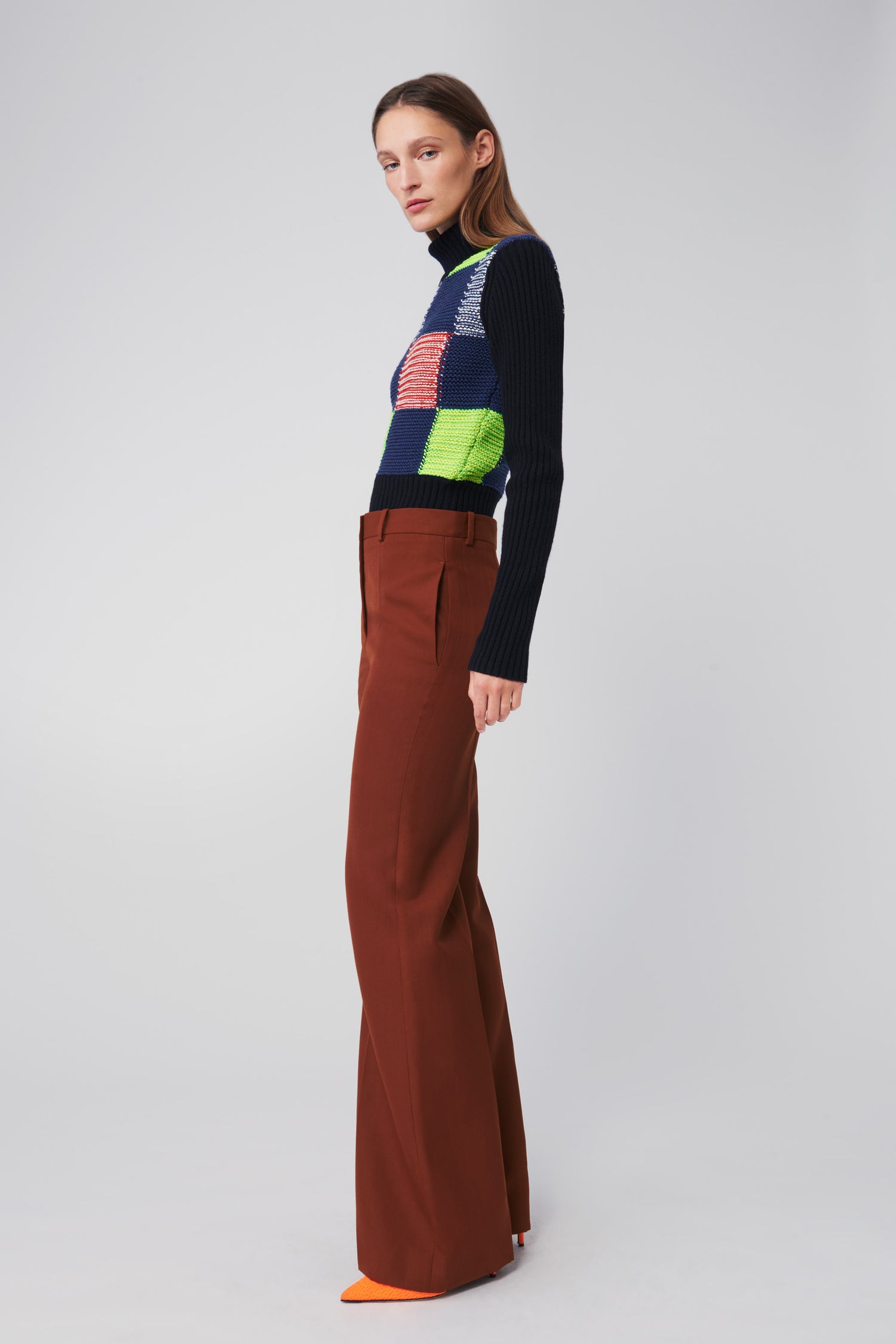 A person stands against a plain background, wearing a Victoria Beckham Patchwork Polo Neck Jumper in Multicolour, paired with brown wide-leg trousers and vibrant orange shoes.