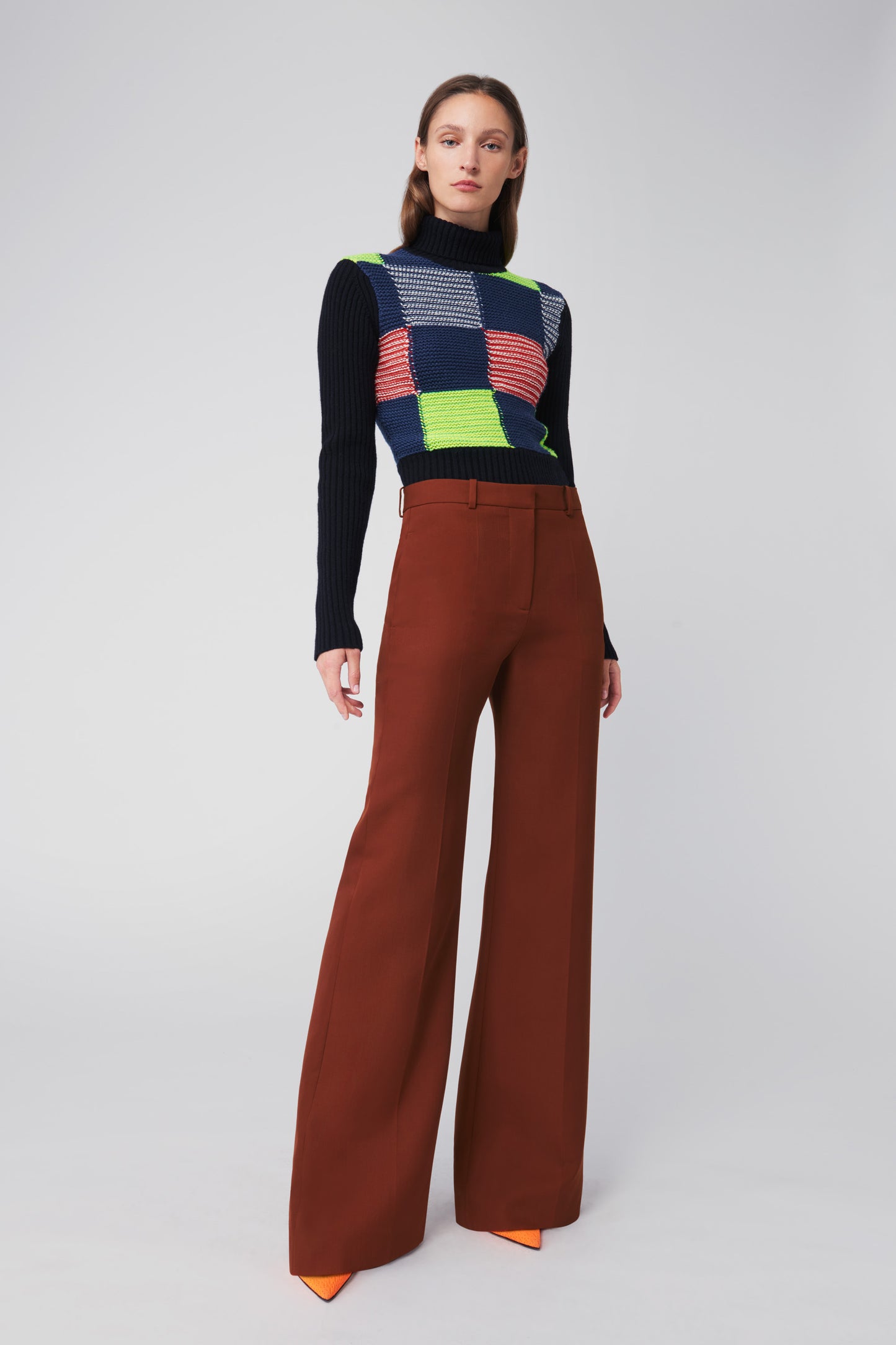 A person stands against a plain background wearing the Victoria Beckham Patchwork Polo Neck Jumper in Multicolour, complemented by brown wide-leg pants and orange shoes.
