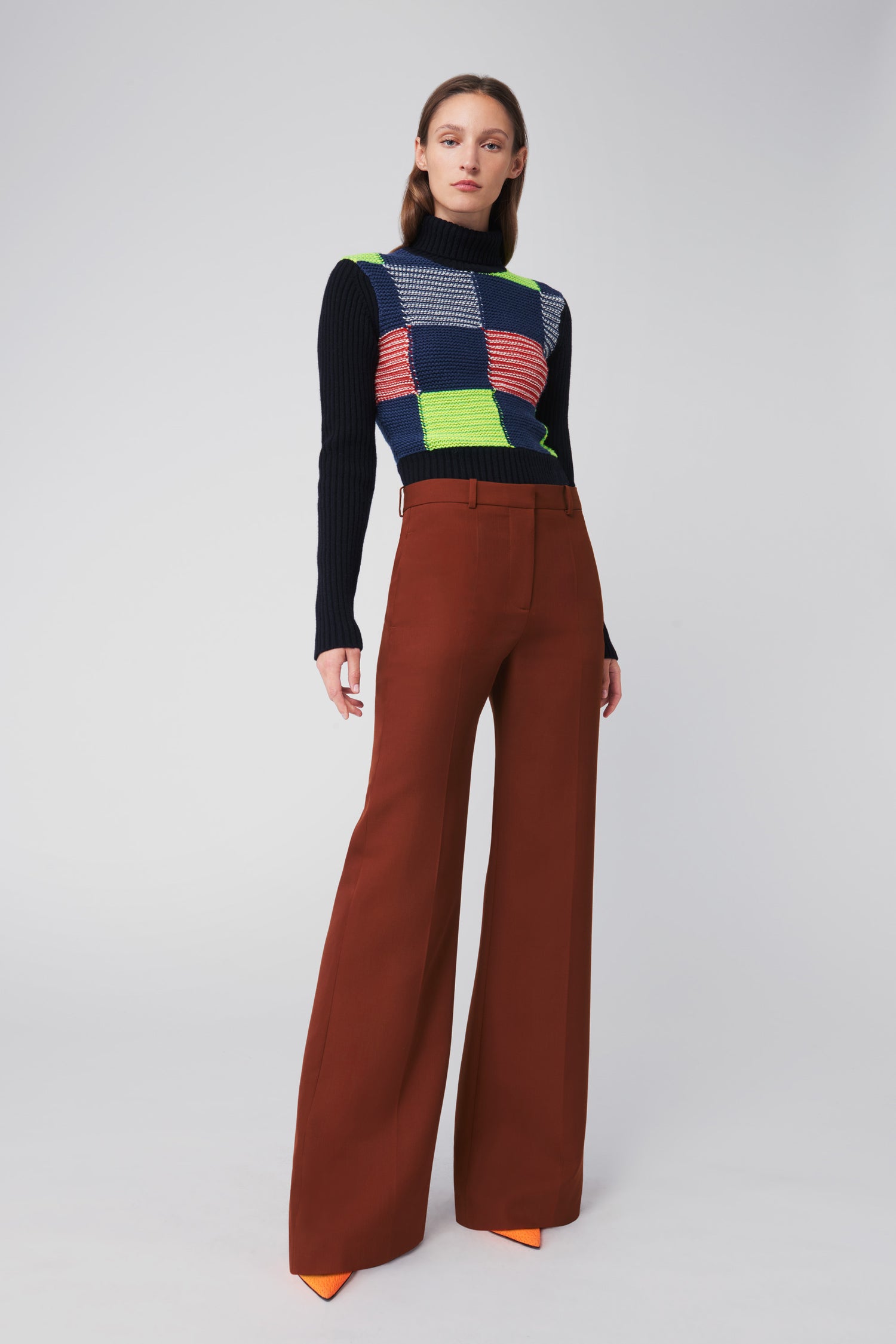 A person stands against a plain background wearing the Victoria Beckham Patchwork Polo Neck Jumper in Multicolour, complemented by brown wide-leg pants and orange shoes.