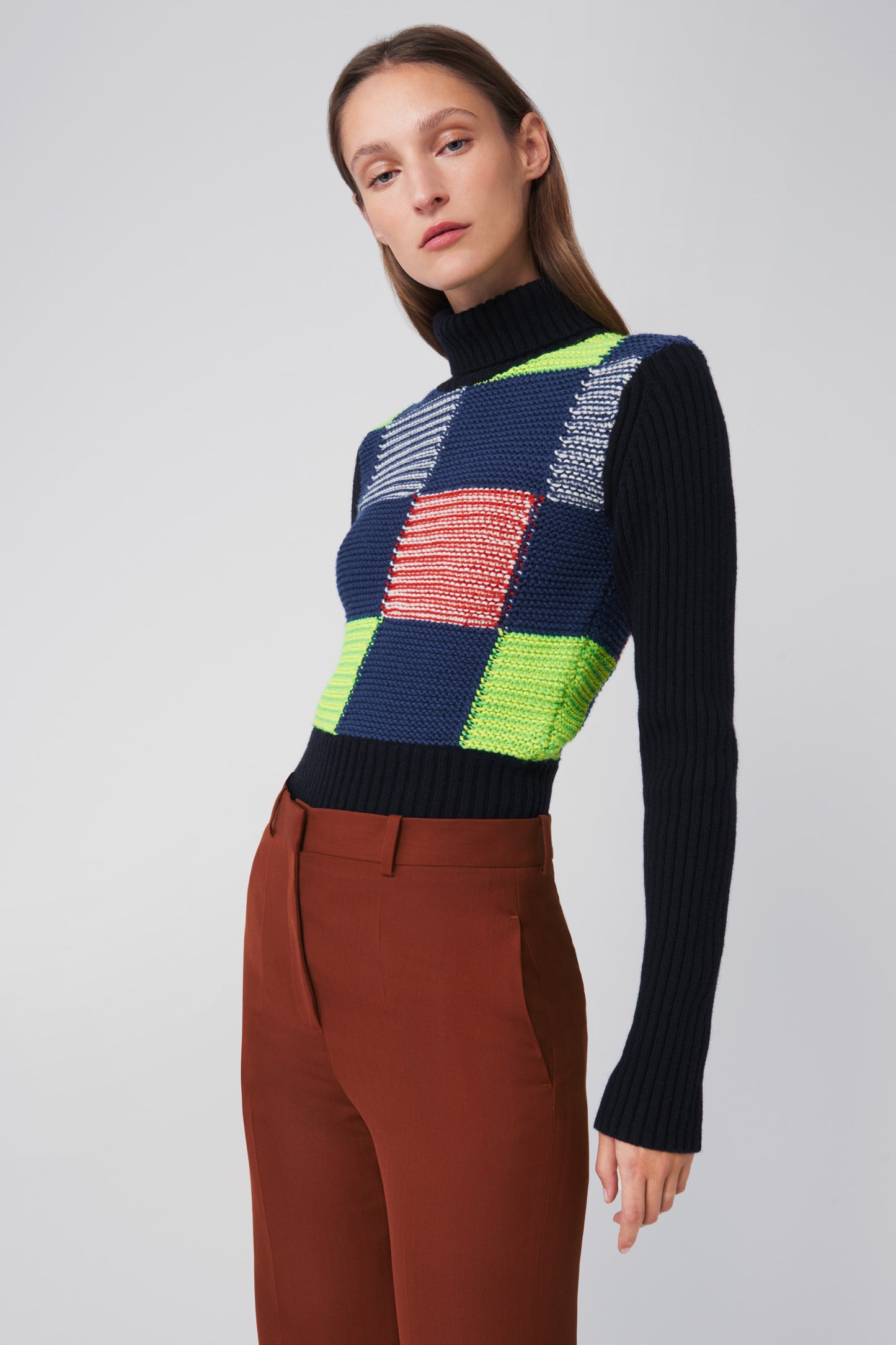 A person is wearing a Victoria Beckham Patchwork Polo Neck Jumper in Multicolour and rust-colored pants, standing against a plain background.