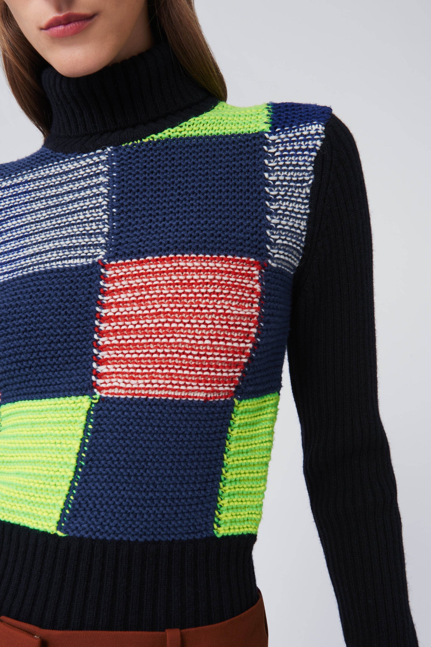 Individual dressed in a Victoria Beckham Patchwork Polo Neck Jumper, featuring multicolored squares of navy, green, red, and white.