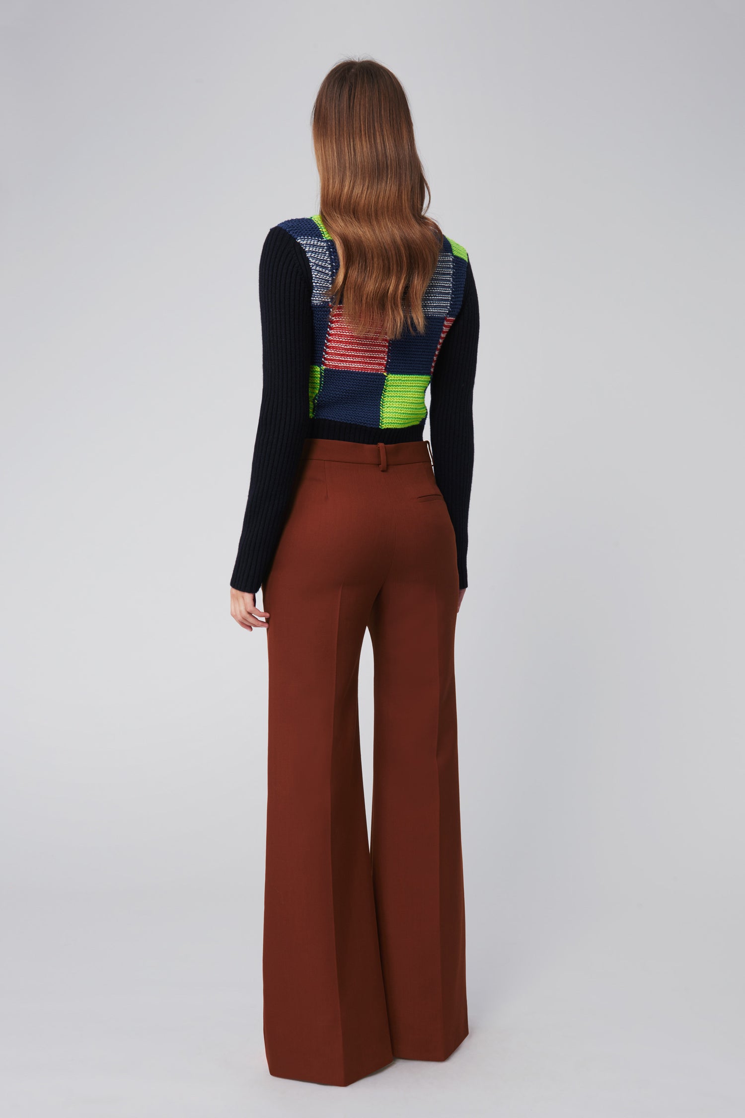 A person with long hair faces away from the camera, wearing the vibrant Victoria Beckham Patchwork Polo Neck Jumper in Multicolour paired with brown wide-leg pants.