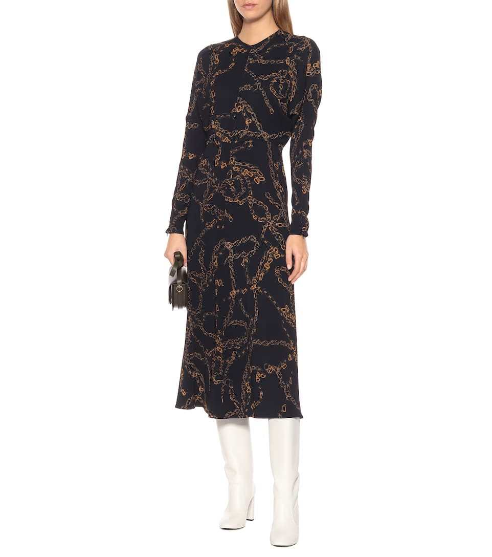 A woman wearing a Victoria Beckham UK Long Sleeve Dolman Midi with a chain pattern, holding a small handbag, and white knee-high boots—reminiscent of Lindsay Broome's signature style.